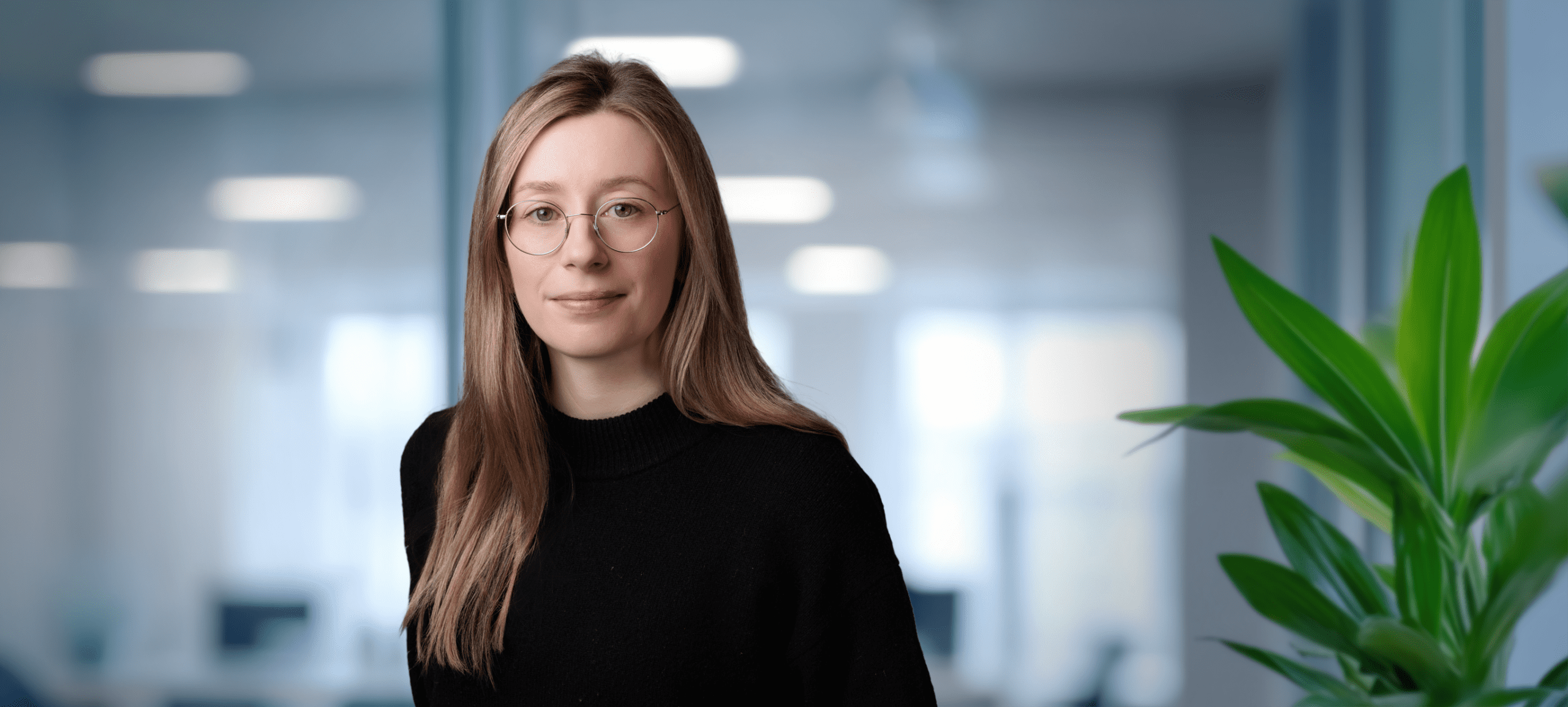 Headshot of a woman with light brown hair and round glasses, wearing a black sweater, against a blurred office background with a green plant.