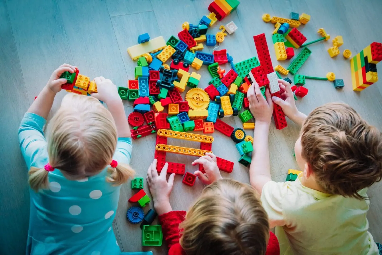 Three children playing with a large pile of colorful building blocks on a wooden floor, seen from an overhead perspective. Their hands are busy connecting various red, blue, green, and yellow blocks.