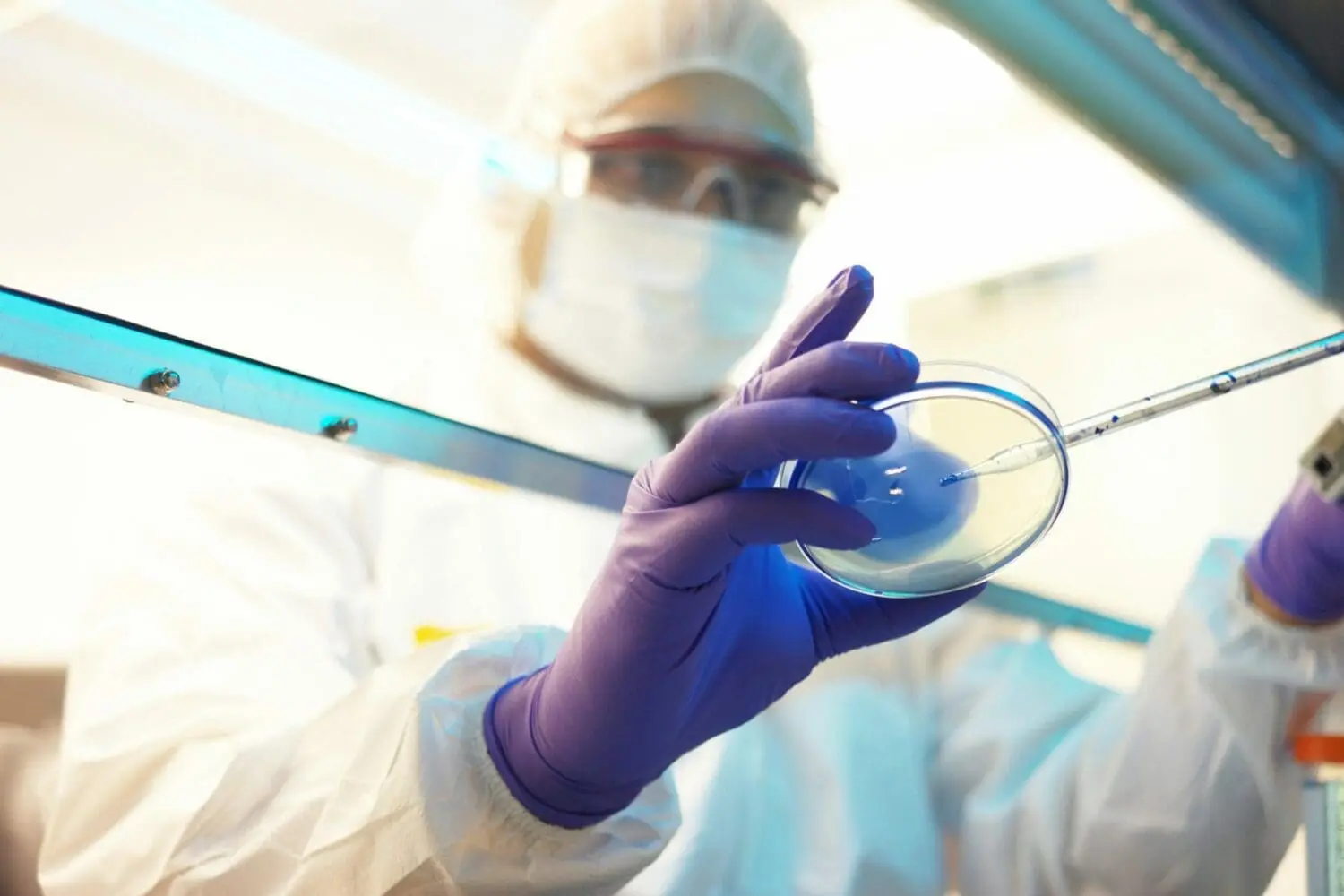 Adherent (formerly Compliance & Risks). A scientist in a white hazmat suit, face mask, safety goggles, and purple gloves, carefully pipetting liquid into a petri dish.