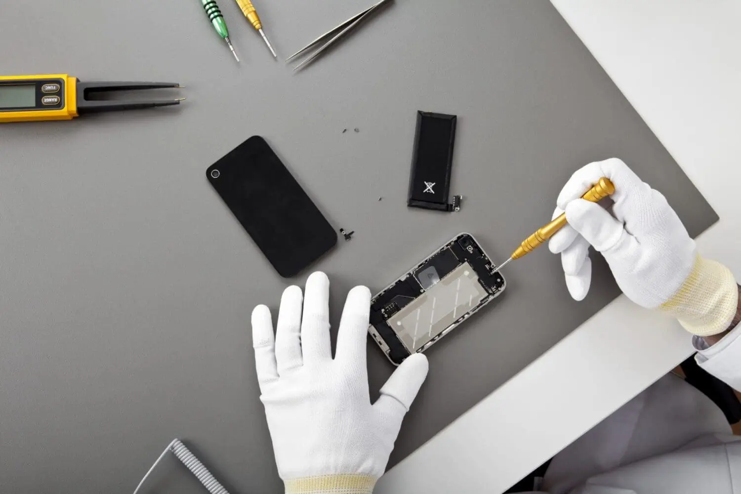 Adherent (formerly Compliance & Risks). Overhead view of gloved hands repairing a smartphone on a gray surface, with tools and disassembled phone parts scattered around.