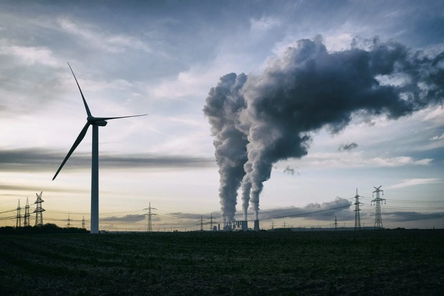 Adherent (formerly Compliance & Risks). A wind turbine on the left, with a power plant emitting dark smoke from multiple stacks on the right, under a cloudy sky. Electricity pylons are visible across the landscape.