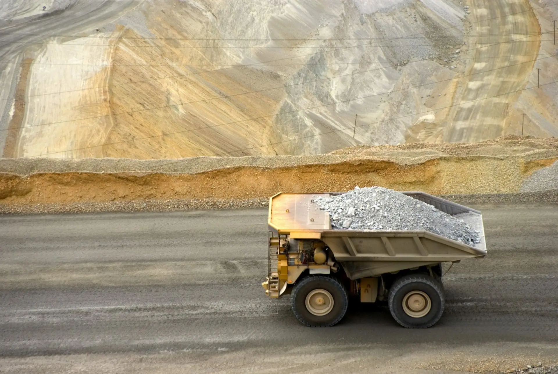 Adherent (formerly Compliance & Risks). A large yellow mining dump truck, full of grey rocks, drives on a dirt road in front of a vast terraced mine wall.