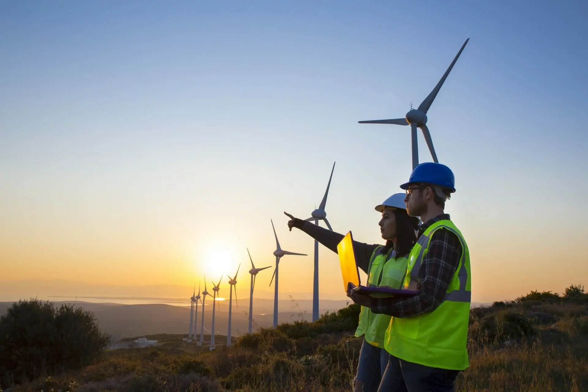 Adherent (formerly Compliance & Risks). Two engineers in hard hats and high-visibility vests stand on a hill, overlooking a wind farm at sunset. One points towards the horizon while holding a laptop.
