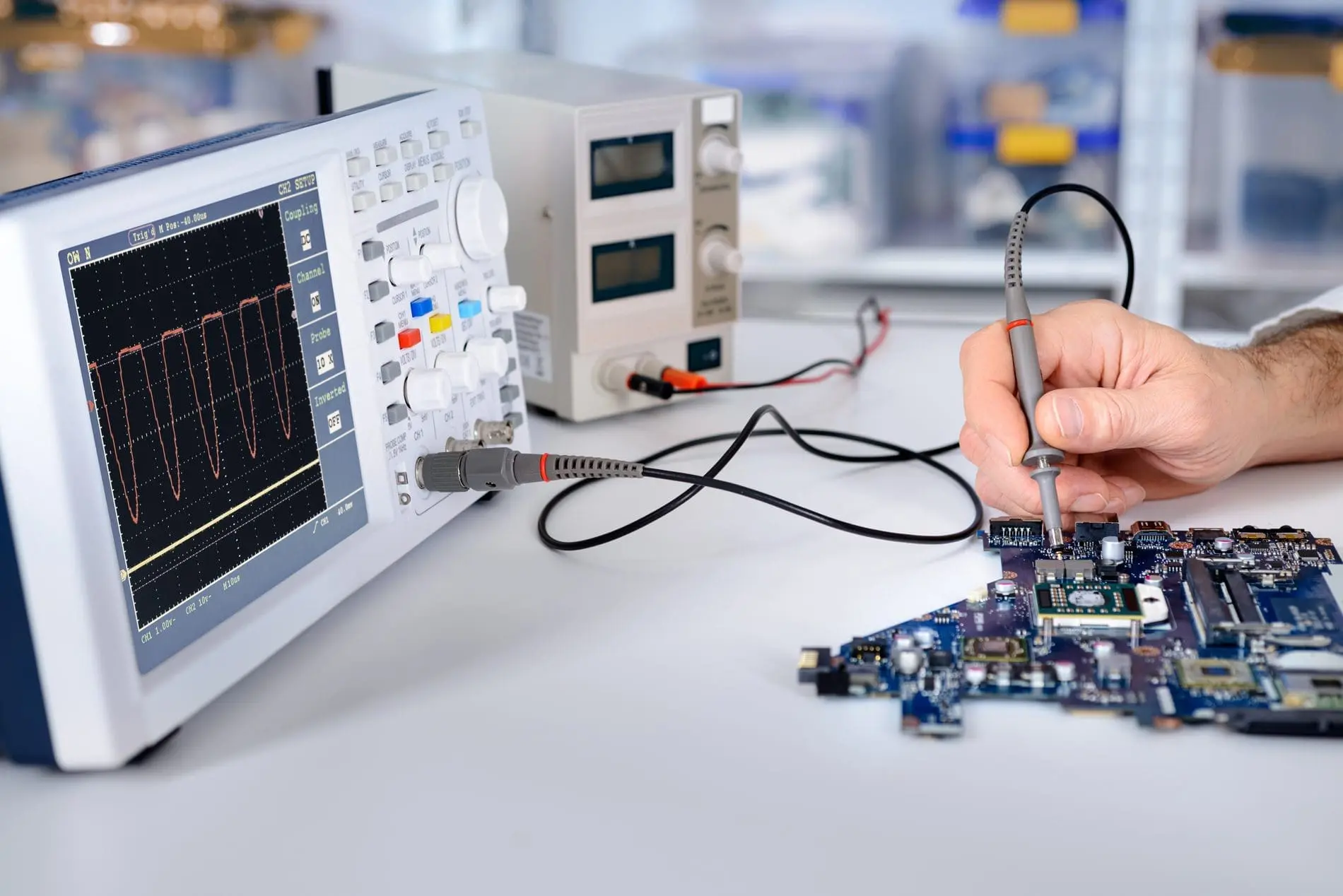 A hand uses a probe to test a blue circuit board next to an oscilloscope displaying a red waveform.