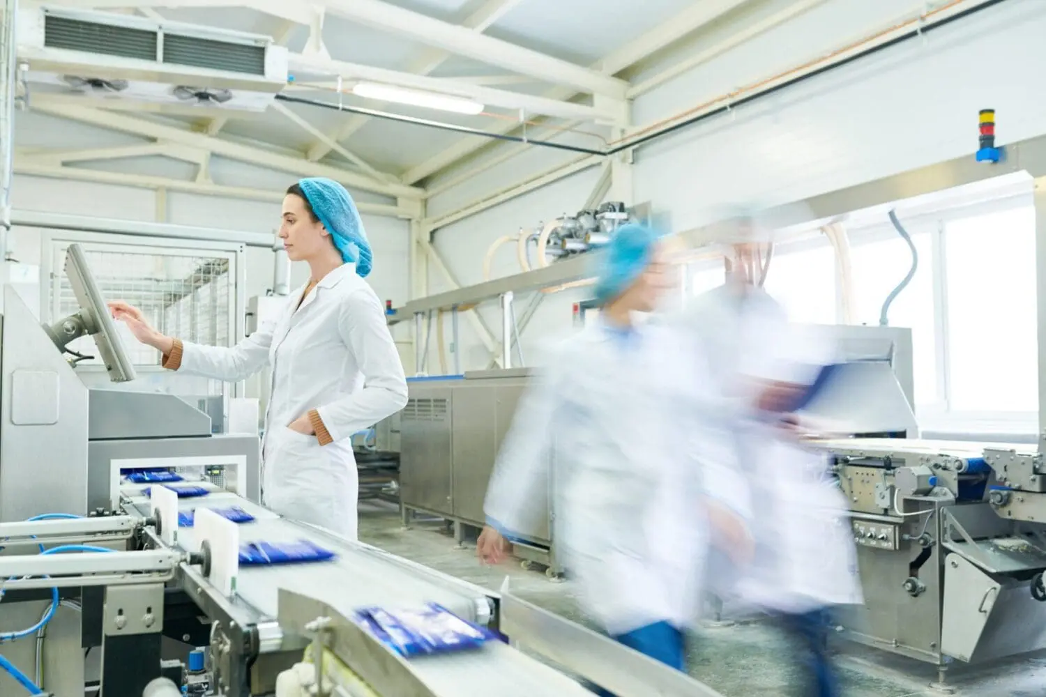 Adherent (formerly Compliance & Risks). A woman in a lab coat and hairnet operates a touchscreen control panel next to a conveyor belt carrying packaged goods in a factory.