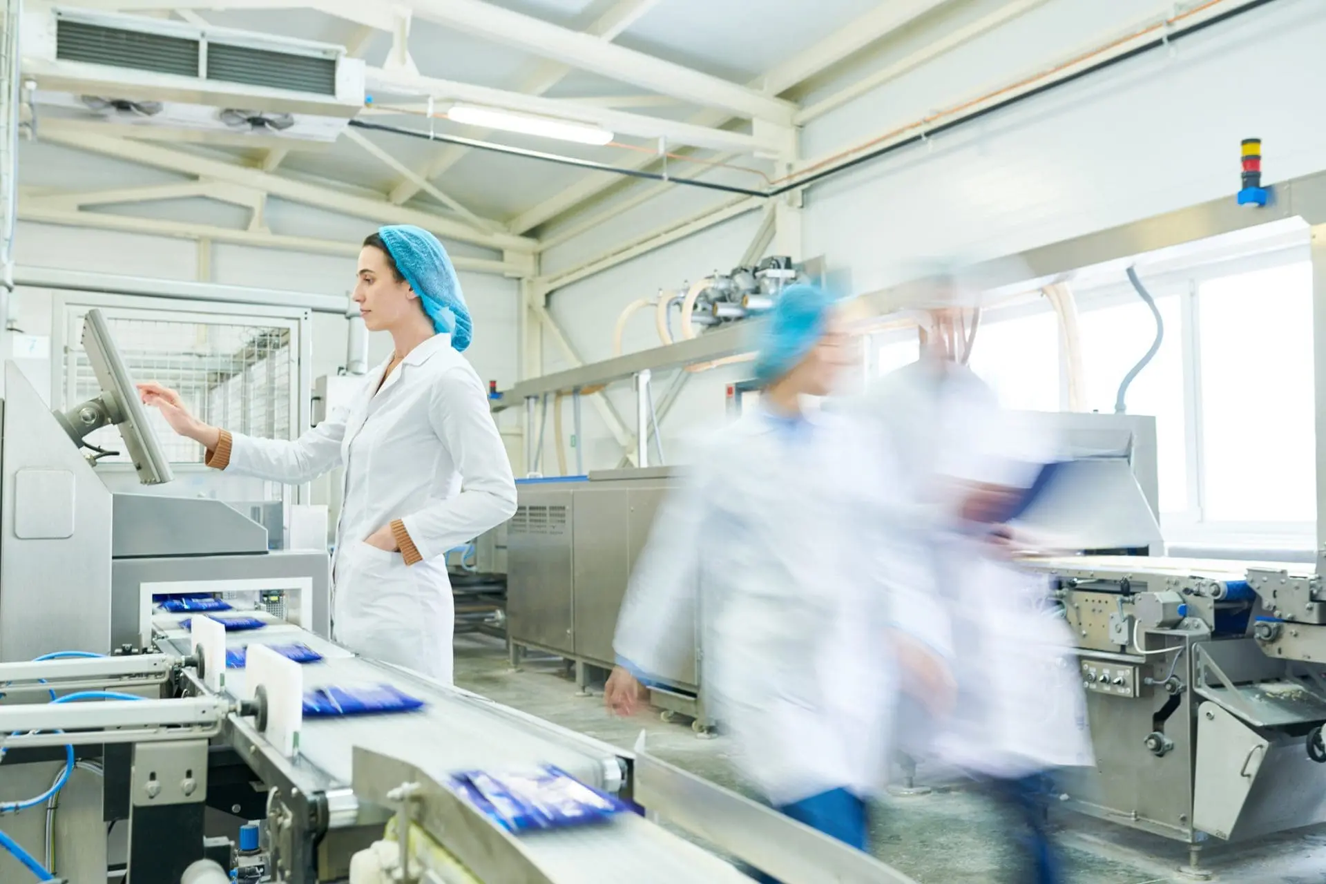 Adherent (formerly Compliance & Risks). A woman in a lab coat and hairnet operates a touchscreen control panel next to a conveyor belt carrying packaged goods in a factory.