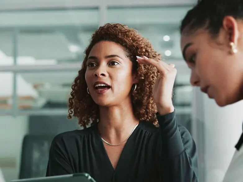 A woman with curly brown hair speaks and gestures with her hand, wearing a dark top and silver necklace, while another woman is partially visible beside her in an office setting.
