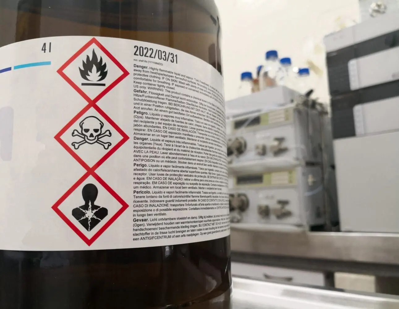 Adherent (formerly Compliance & Risks). A brown chemical bottle with a white label showing GHS pictograms for flammability, toxicity, and health hazard, along with warning text and a shelf life date of 2022/03/31.
