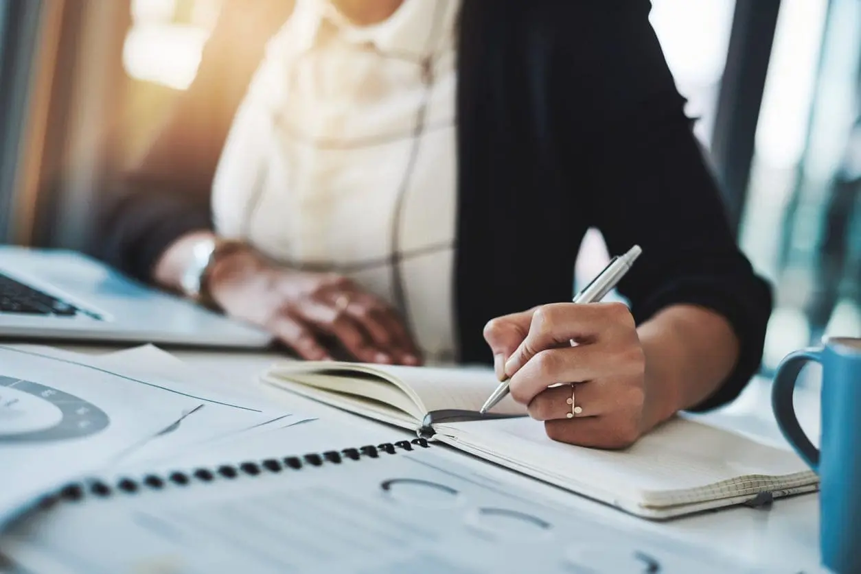 Adherent (formerly Compliance & Risks). A person's hands writing in an open notebook with a silver pen, surrounded by a laptop, papers with charts, and a blue mug on a desk.