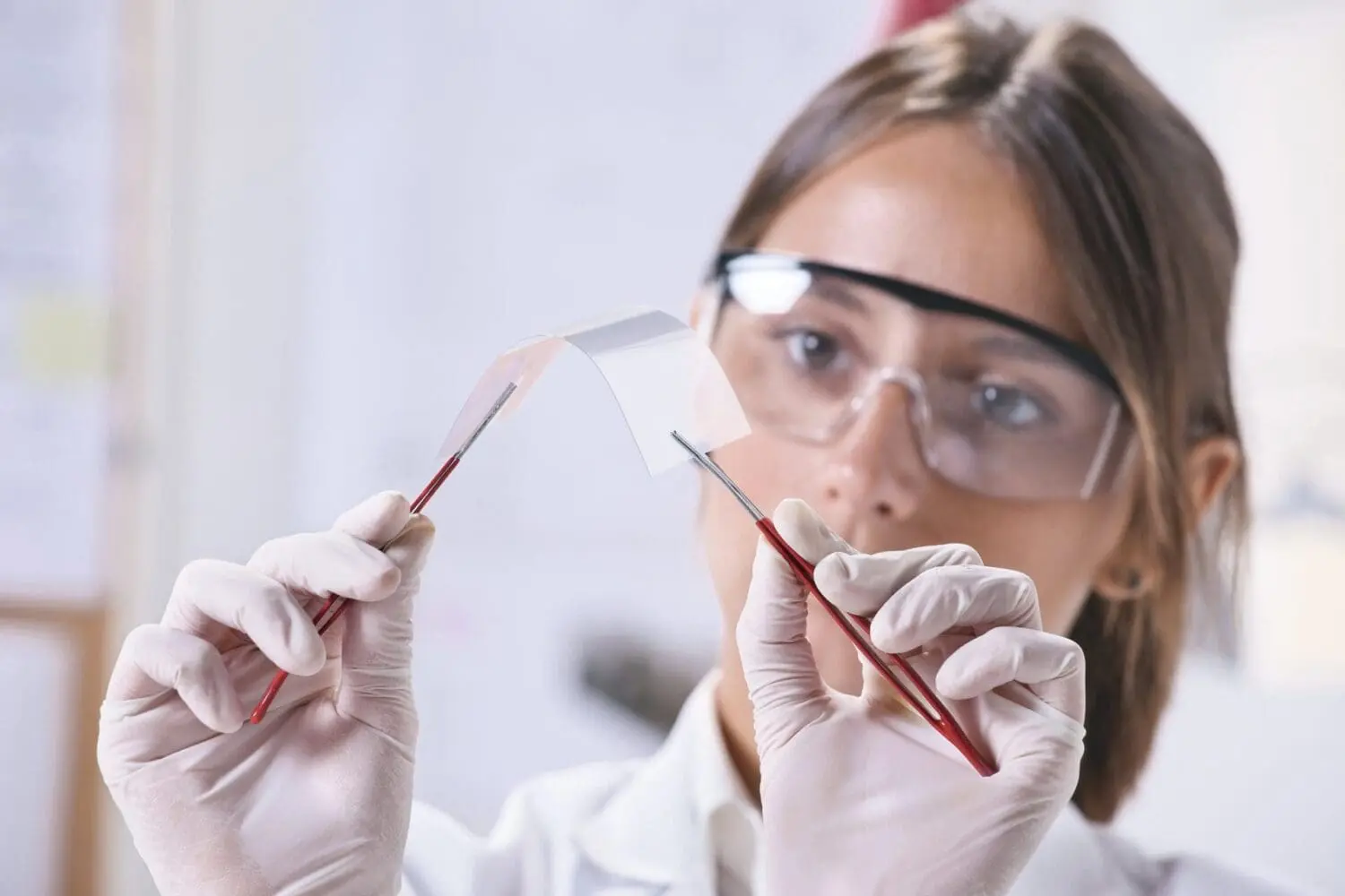 Adherent (formerly Compliance & Risks). A scientist wearing safety glasses and white gloves holds a flexible, translucent material with two red-handled tweezers.