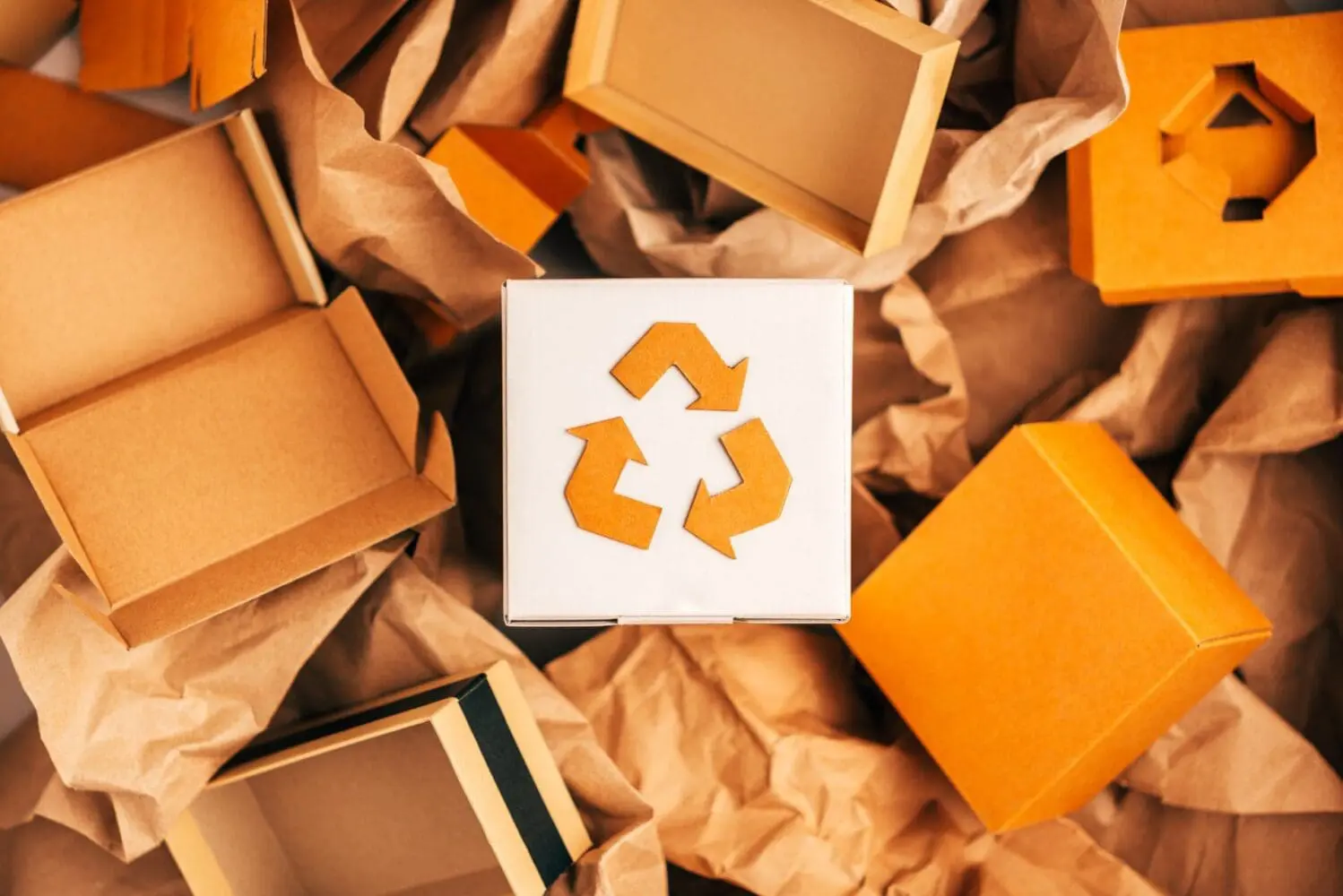 Adherent (formerly Compliance & Risks). Overhead view of various brown and orange cardboard boxes and crumpled packing paper, with a white box in the center displaying an orange recycling symbol.