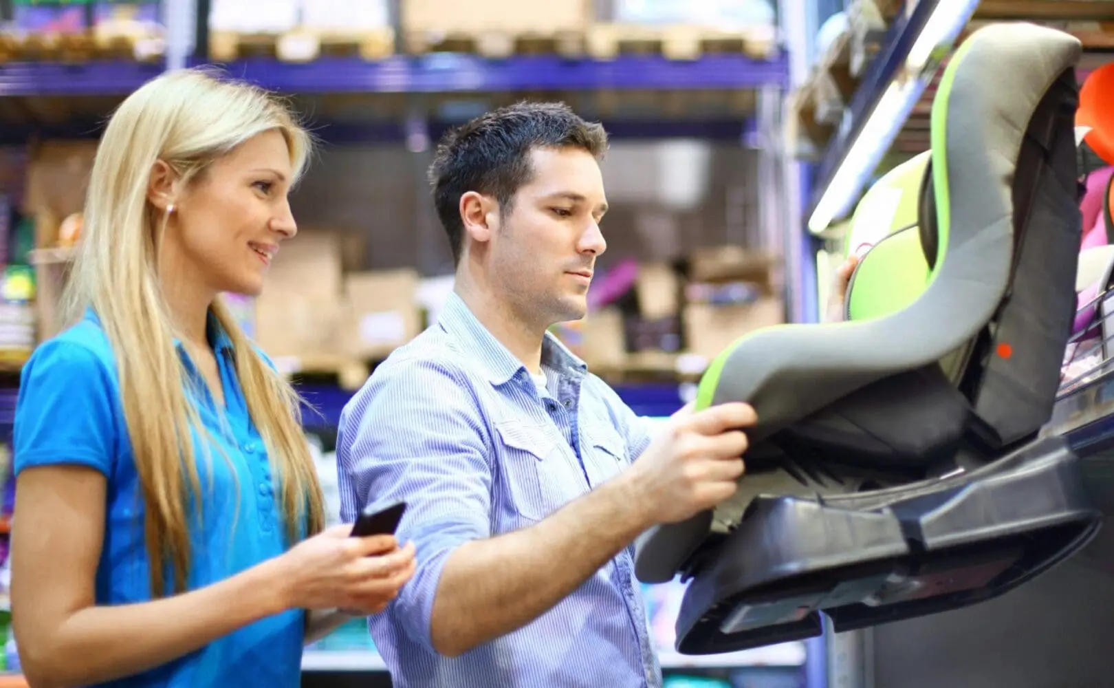 Adherent (formerly Compliance & Risks). A man and a woman in a store aisle, looking at a grey and lime green baby car seat. The woman holds a phone and smiles, while the man examines the car seat.