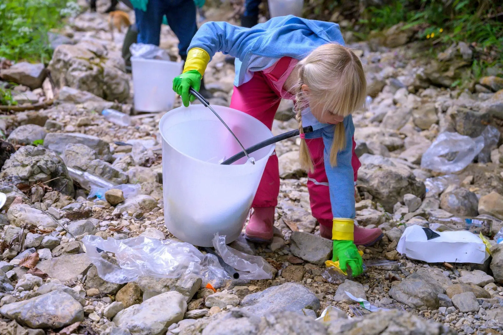 A young girl in a blue hoodie and pink pants, wearing green gloves, uses a grabber tool to pick up trash and place it into a white bucket on a rocky, littered ground.