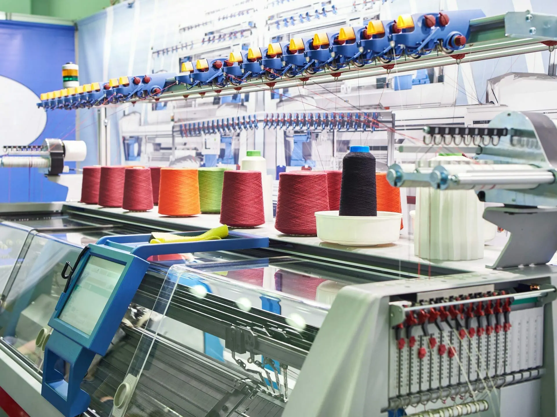 Industrial knitting machine with colorful spools of thread in red, orange, green, and black, and a digital control panel.