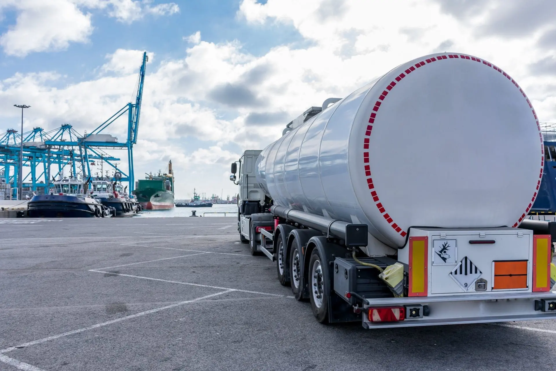 A white tanker truck parked at a port with blue gantry cranes, tugboats, and a cargo ship in the background under a cloudy sky.