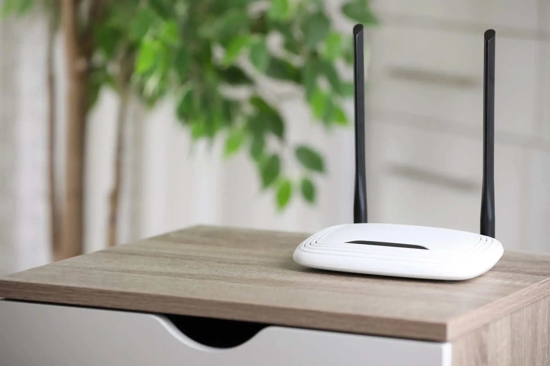 A white Wi-Fi router with two black antennas on a light brown wooden table, with a blurred green plant and light background.