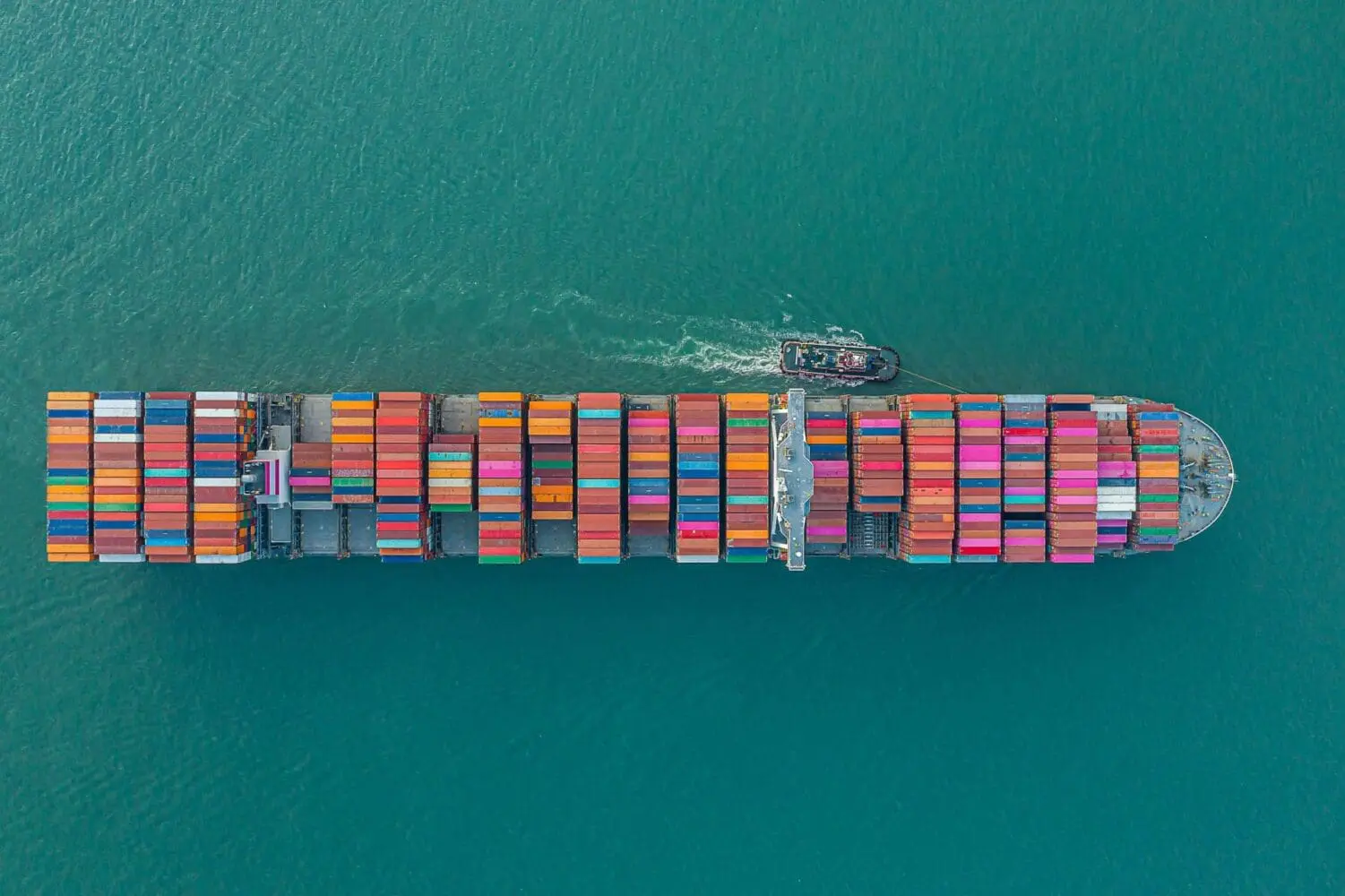 Adherent (formerly Compliance & Risks). Aerial view of a large container ship filled with colorful shipping containers on teal water, with a tugboat at its stern.