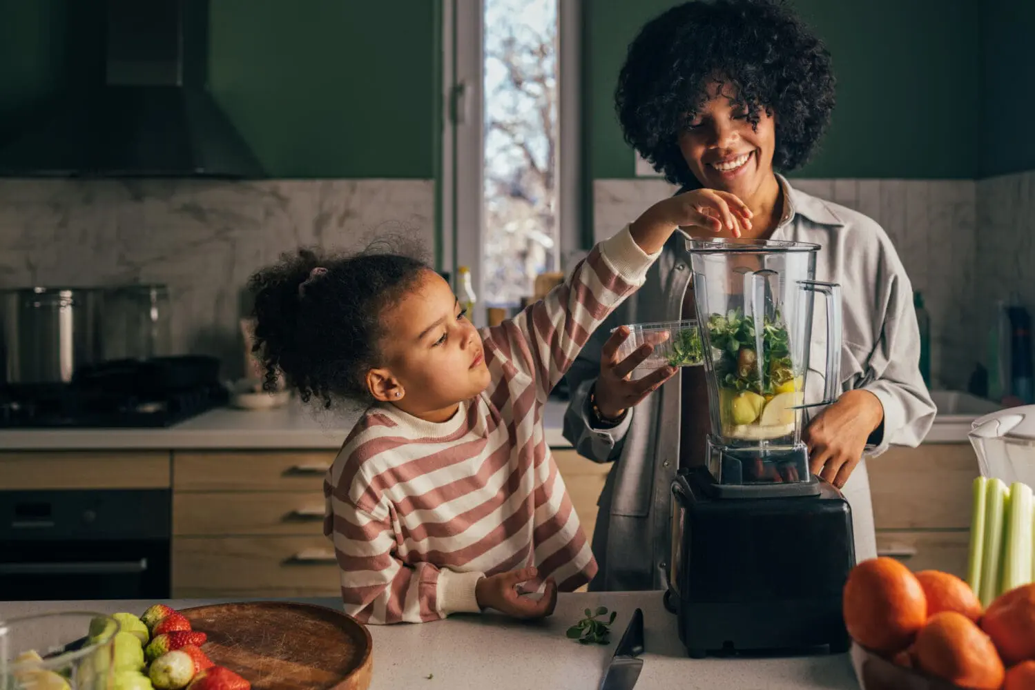 A smiling Black mother and her young daughter making a green smoothie in a kitchen. The daughter is adding greens to the blender, surrounded by fresh fruits like oranges, strawberries, and celery.