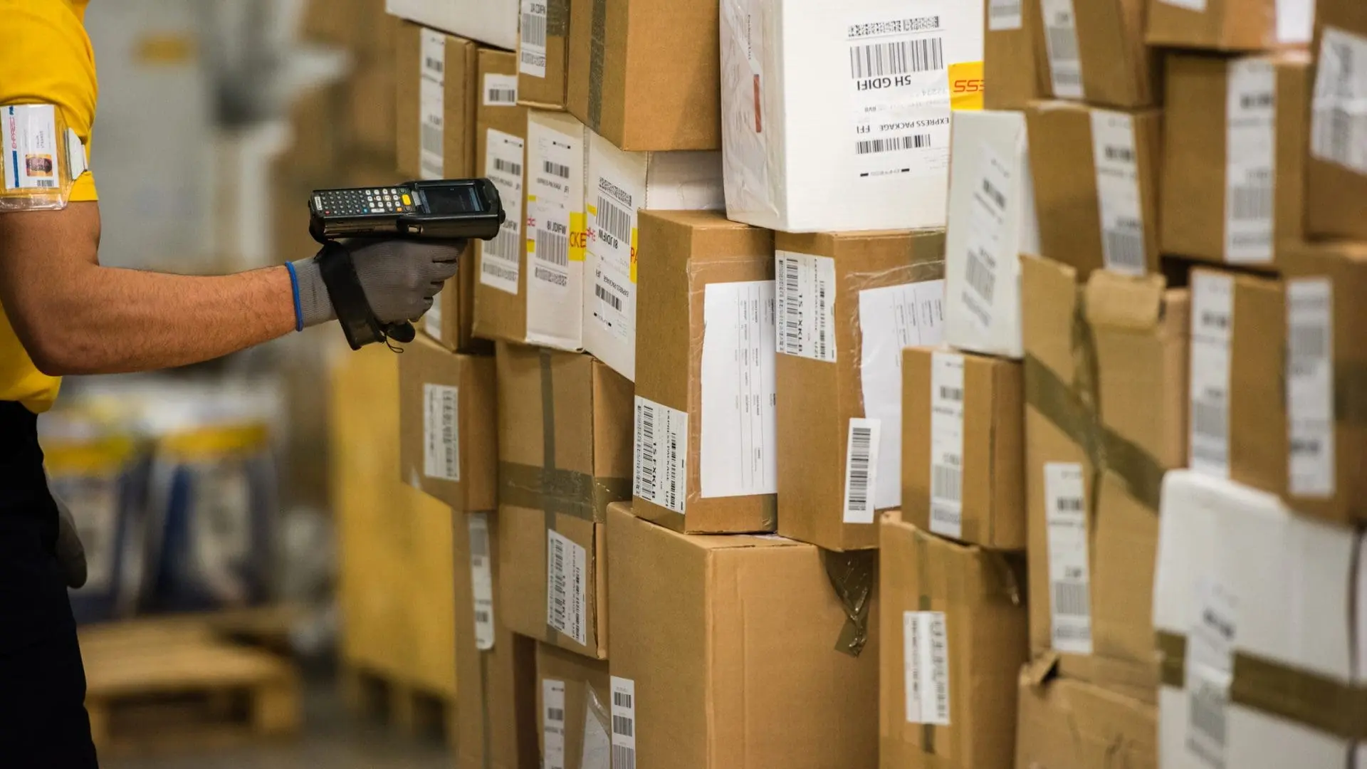 A person in a yellow shirt and grey glove uses a handheld barcode scanner to scan a tall stack of cardboard shipping boxes in a warehouse.