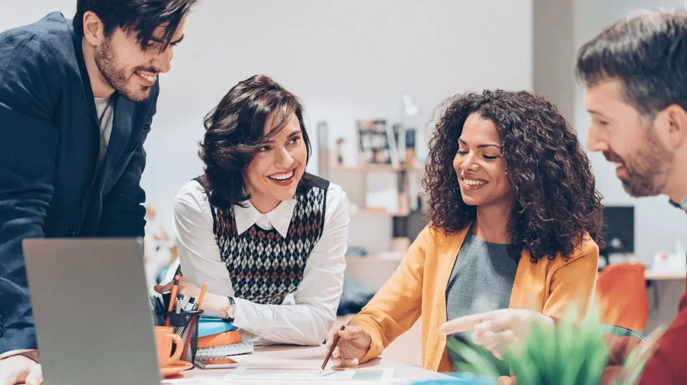 Four smiling colleagues, two men and two women, collaborate around a desk with a laptop and documents in a bright office.