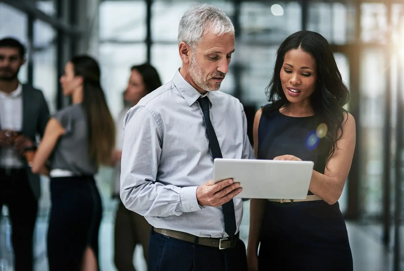 A man and a woman in an office setting look intently at a white tablet, appearing to discuss content on the screen. Other blurred colleagues are in the background.