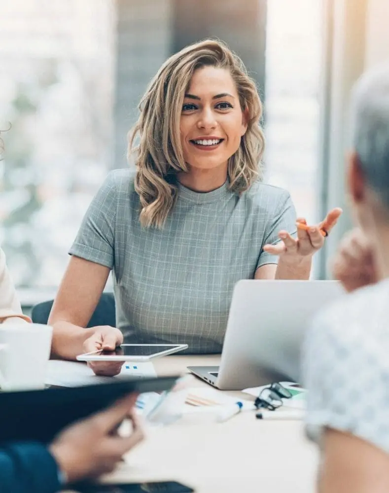 Adherent (formerly Compliance & Risks). Four diverse professionals in a bright office meeting. A blonde woman smiles and gestures while speaking, surrounded by laptops and papers.