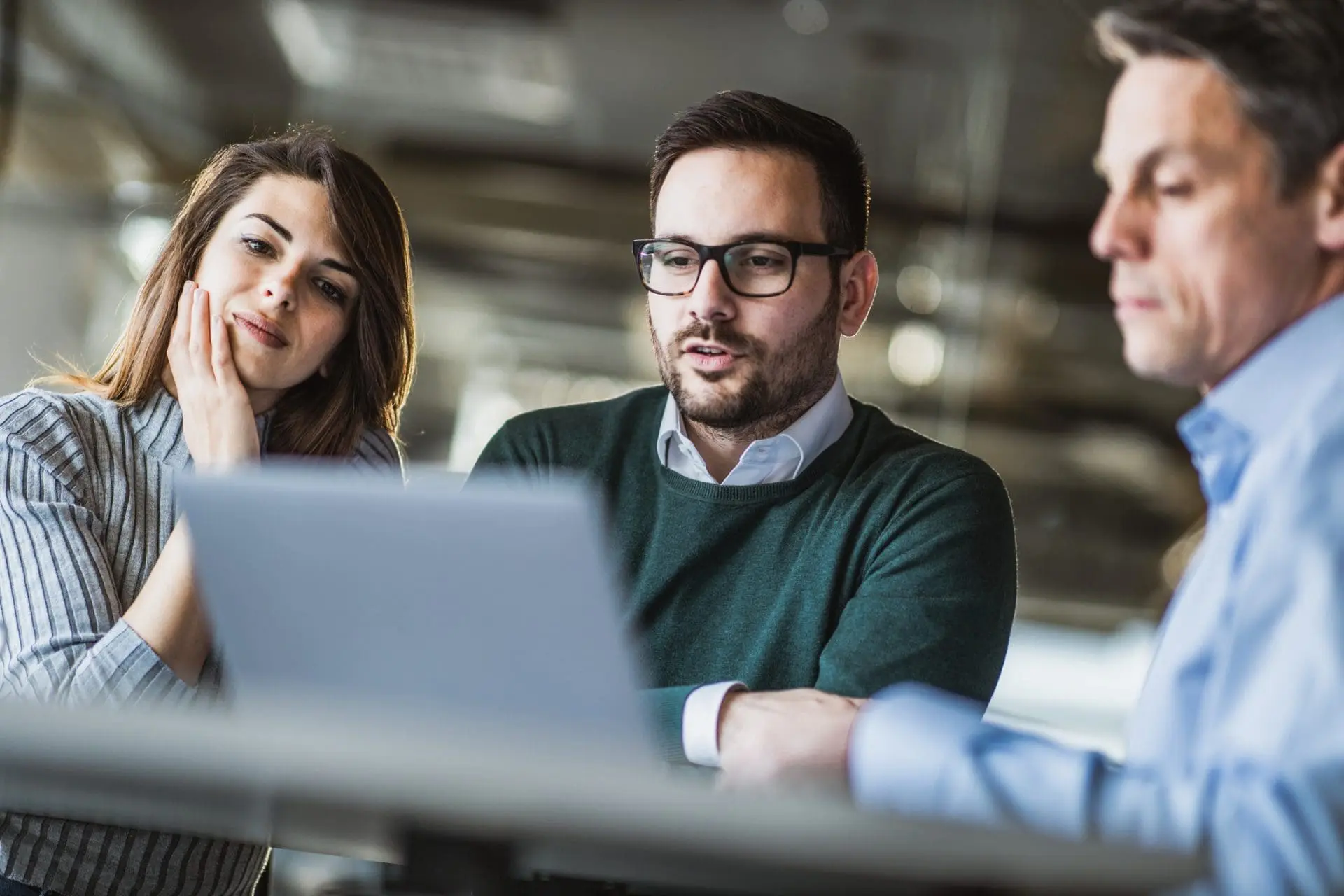 Three diverse colleagues, two men and one woman, intently looking at a laptop screen during a collaborative meeting in an office.