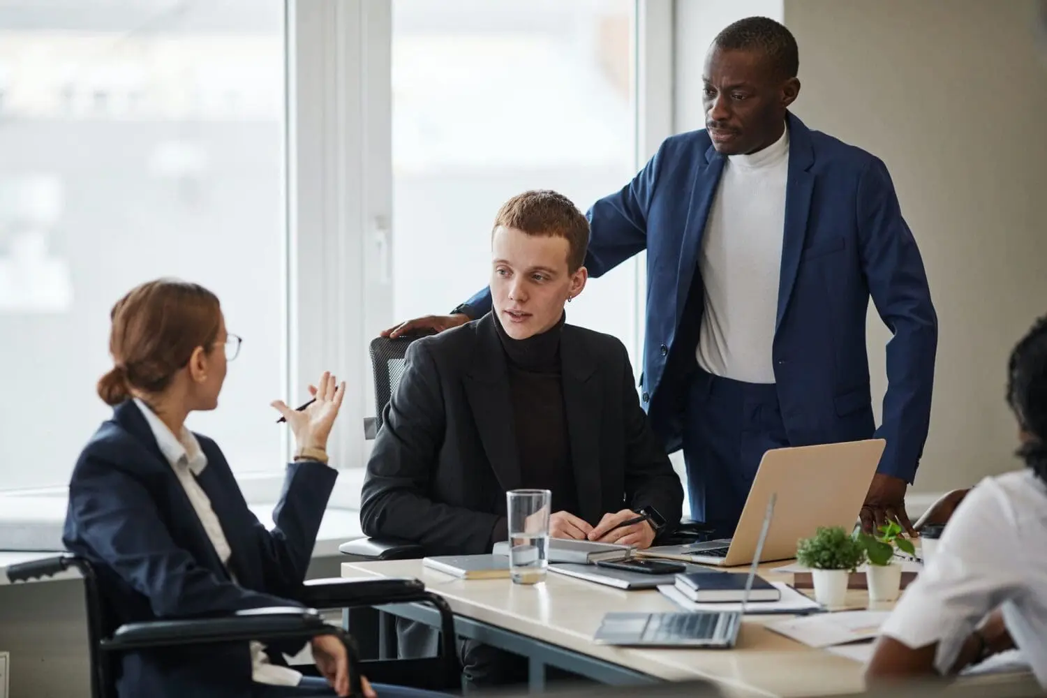 Four professionals, including a woman in a wheelchair, a man in a black suit, and a man in a blue suit, are gathered around a table in an office, engaged in a discussion.