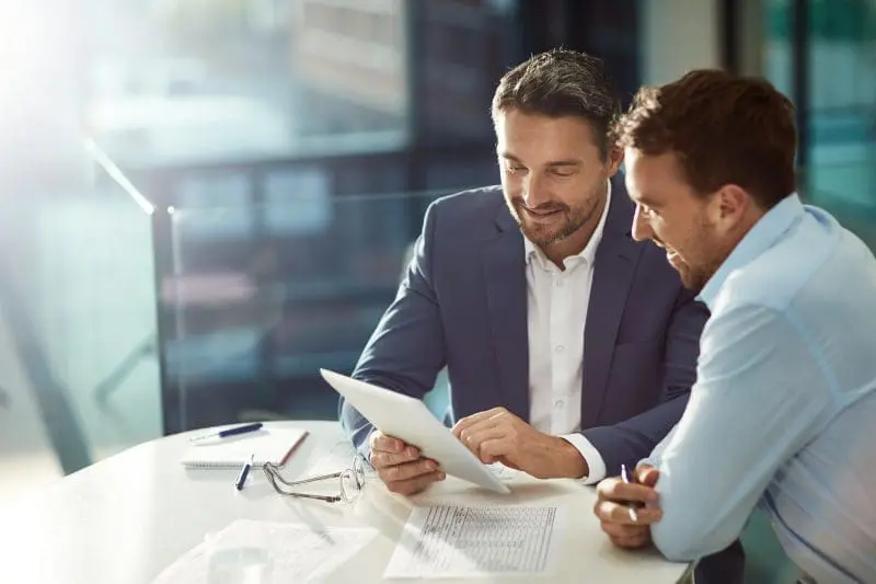 Adherent (formerly Compliance & Risks). Two men in business attire sit at a table, looking intently at a white tablet held by one of them, appearing to discuss its content.
