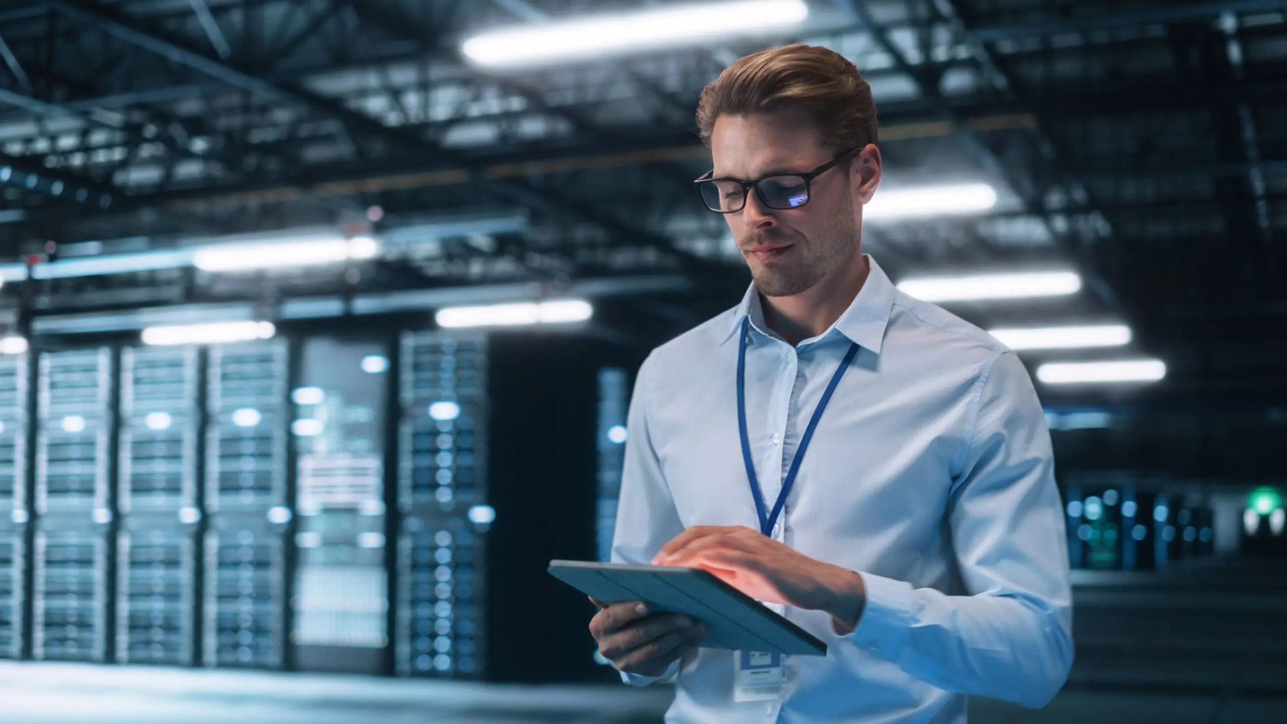 A man in glasses and a light blue shirt uses a tablet in a blurred data center with server racks and bright lights.