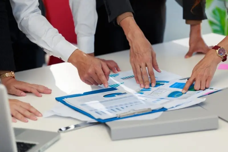 Close-up of multiple hands pointing at and discussing charts and graphs on documents spread across a white table, with a laptop nearby.