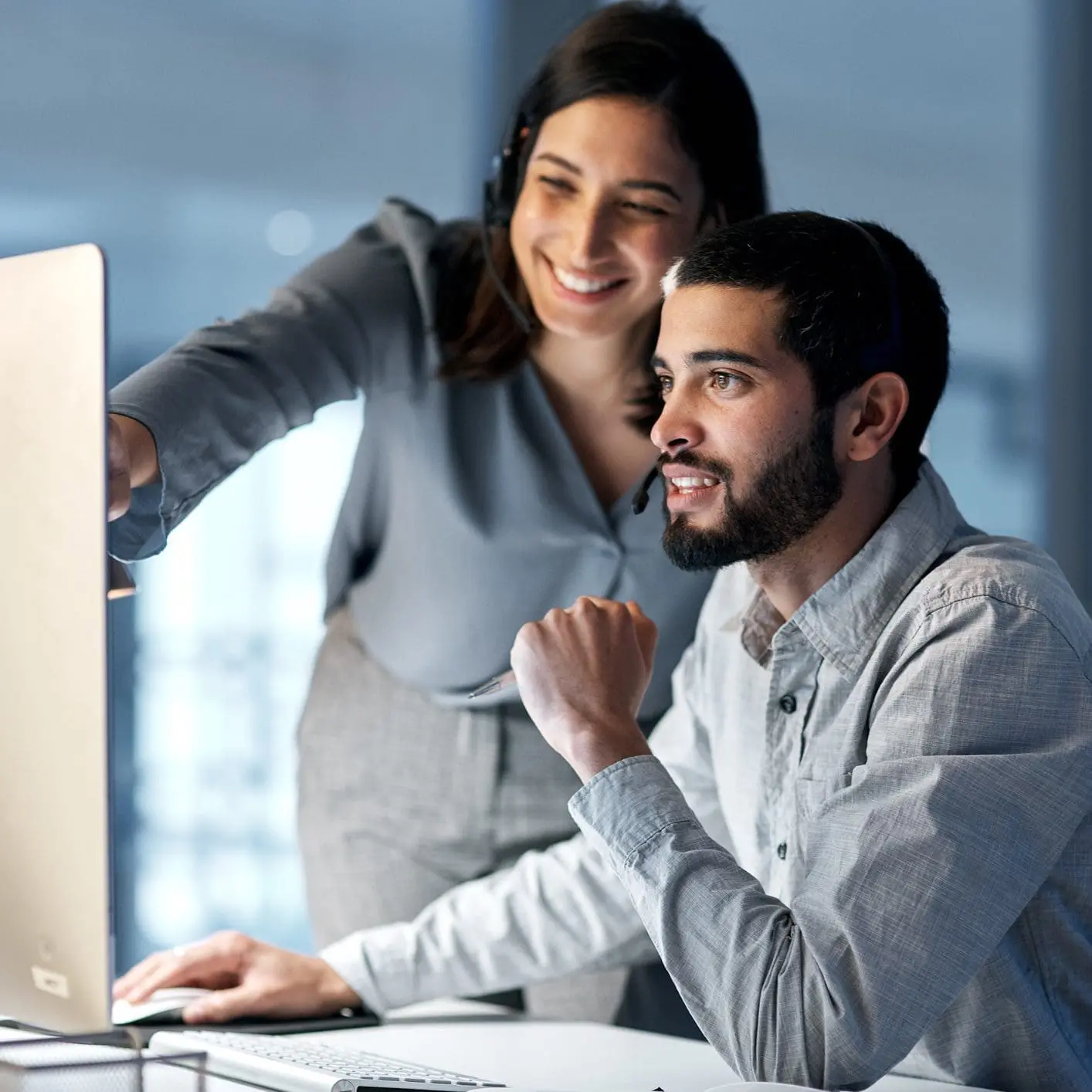 Adherent (formerly Compliance & Risks). Two smiling customer service representatives, a man and a woman, wearing headsets, look at a computer screen together. The woman points at the screen while the man operates the mouse.