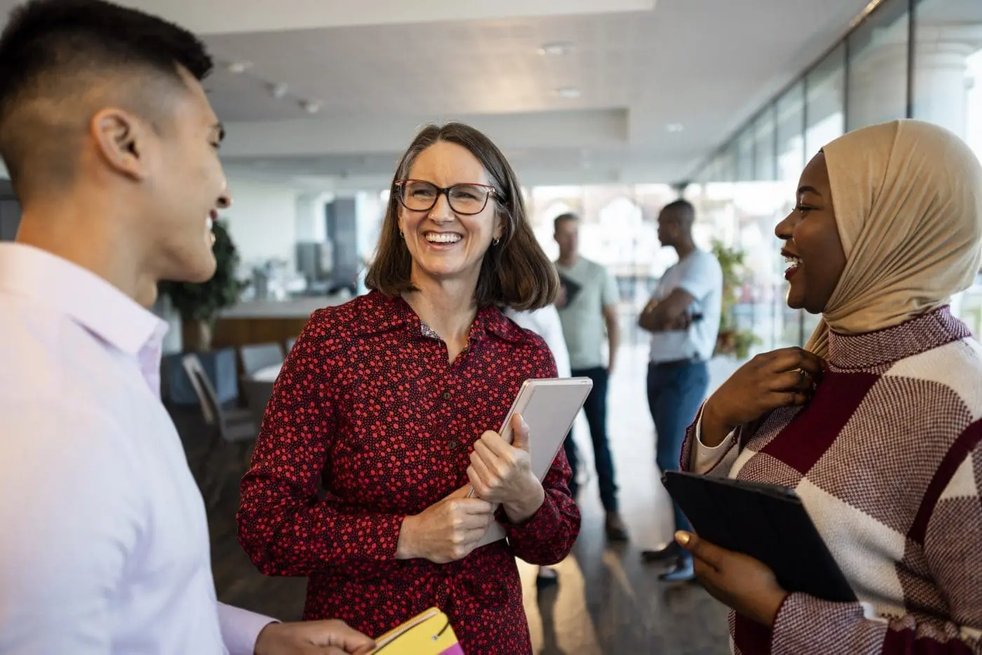 Three smiling, diverse colleagues, two women and one man, engaged in conversation in a modern office. The woman in the center wears glasses and a red patterned shirt, holding a tablet.