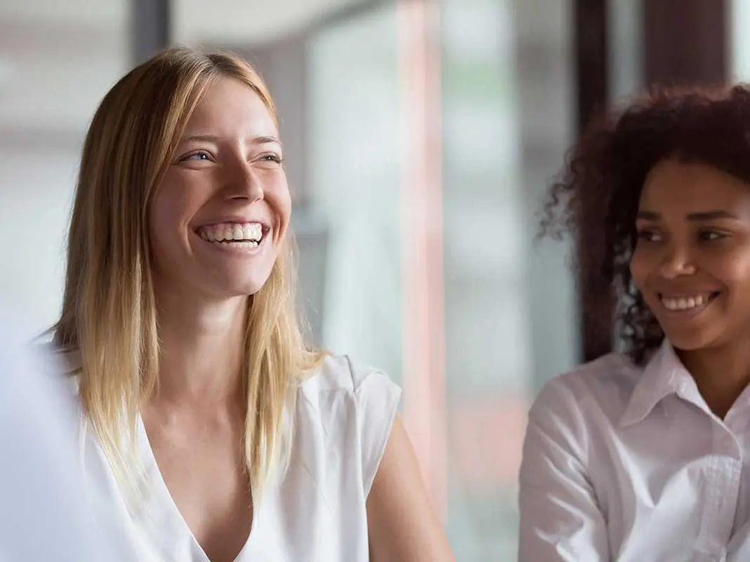 Two women smiling and laughing. The woman in the foreground has blonde hair and is laughing heartily, while the woman behind her with dark curly hair smiles pleasantly.