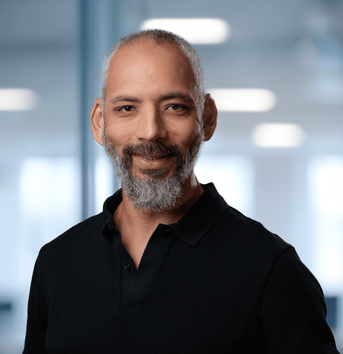 Adherent (formerly Compliance & Risks). Headshot of a smiling man with a salt-and-pepper beard and graying hair, wearing a black polo shirt, in a blurred modern office background with a green plant.