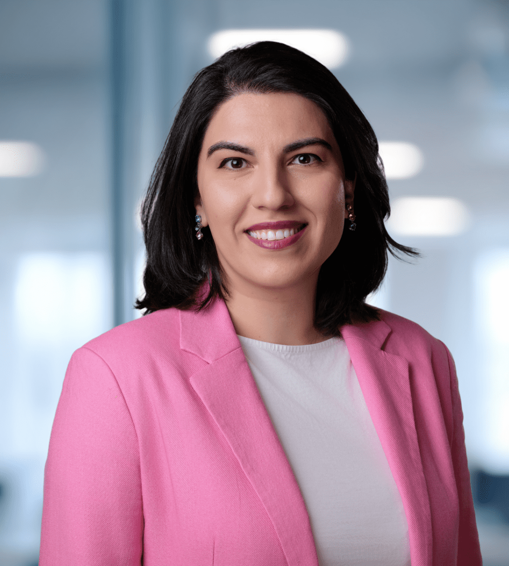 Adherent (formerly Compliance & Risks). A smiling woman with dark hair and a bright pink blazer looks directly at the camera in a blurred modern office environment.