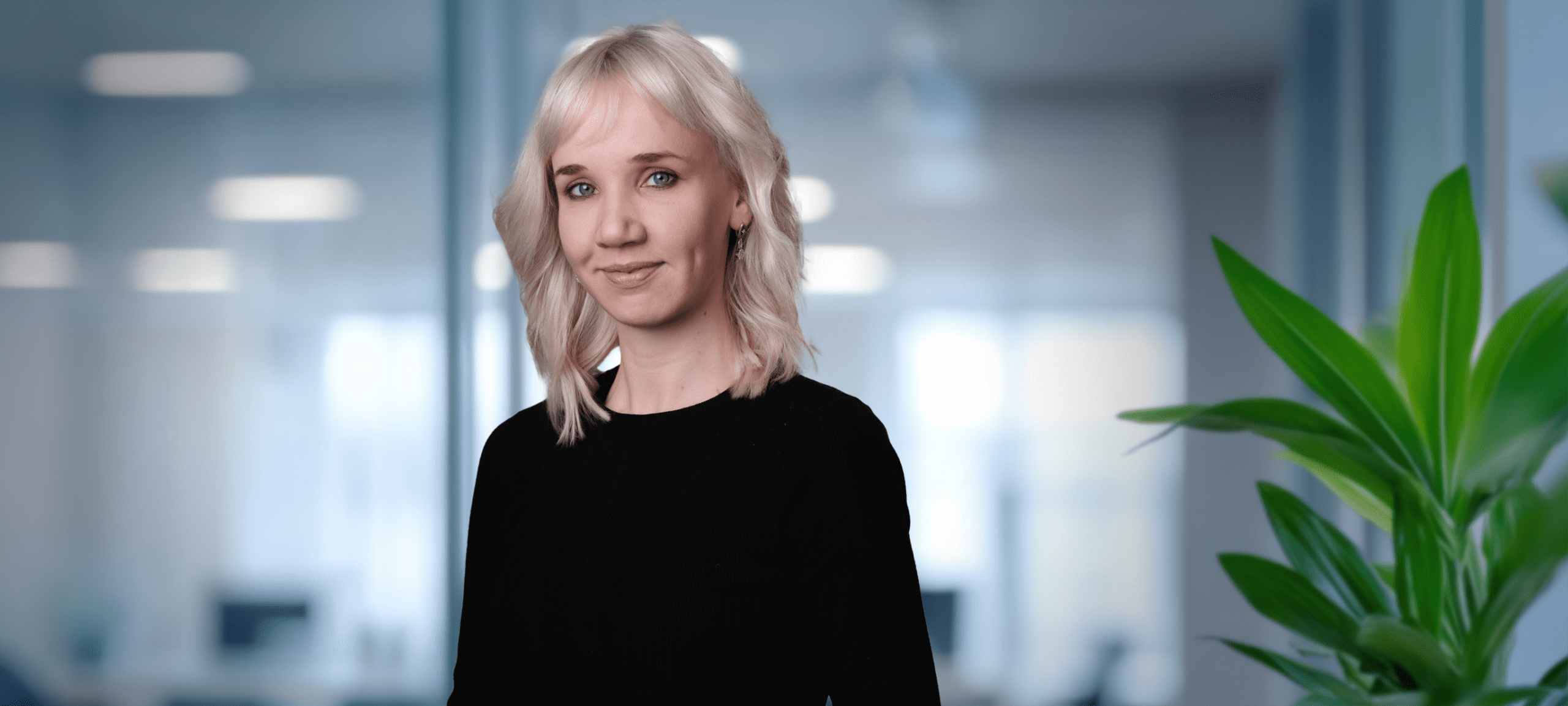 Adherent (formerly Compliance & Risks). A young woman with blonde hair and blue eyes smiles gently in a professional office setting, wearing a black top. A green plant is visible on the right.