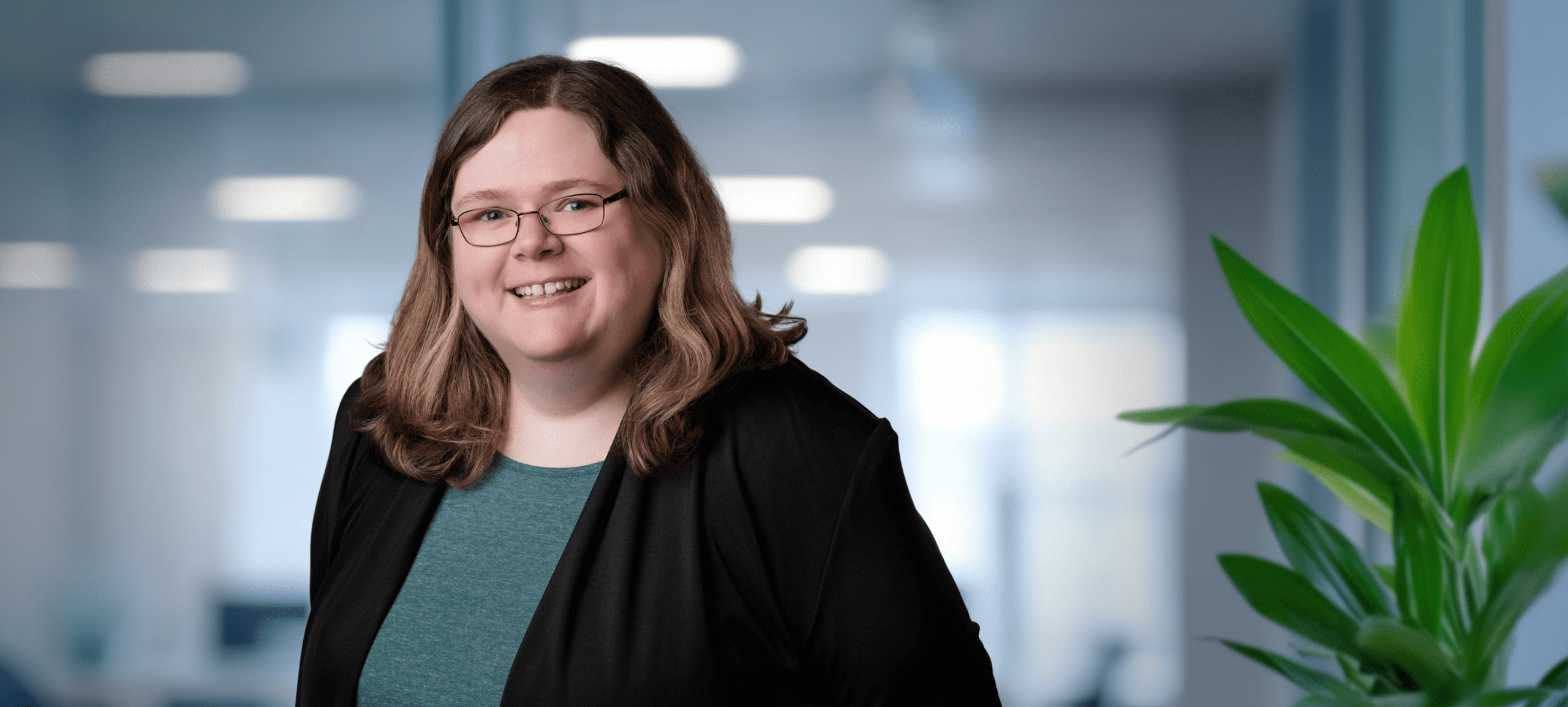Adherent (formerly Compliance & Risks). Headshot of a smiling woman with brown hair and glasses, wearing a teal top and black cardigan, in a blurred office background.