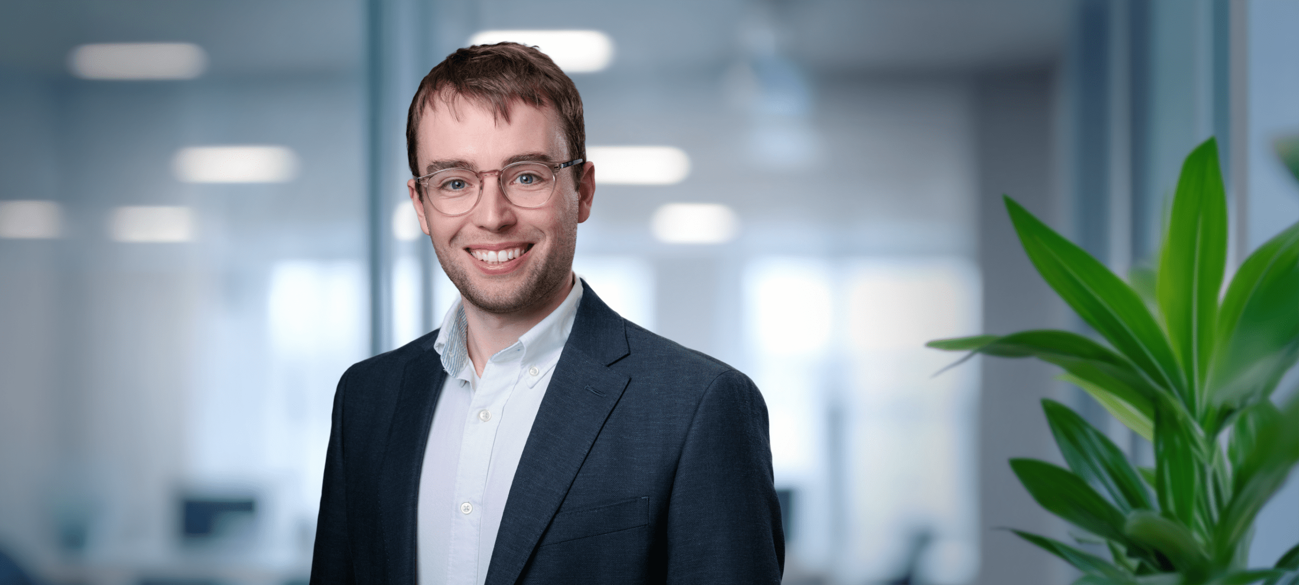 Adherent (formerly Compliance & Risks). Headshot of a smiling young man with light brown hair and glasses, wearing a suit jacket, against a blurred modern office background.