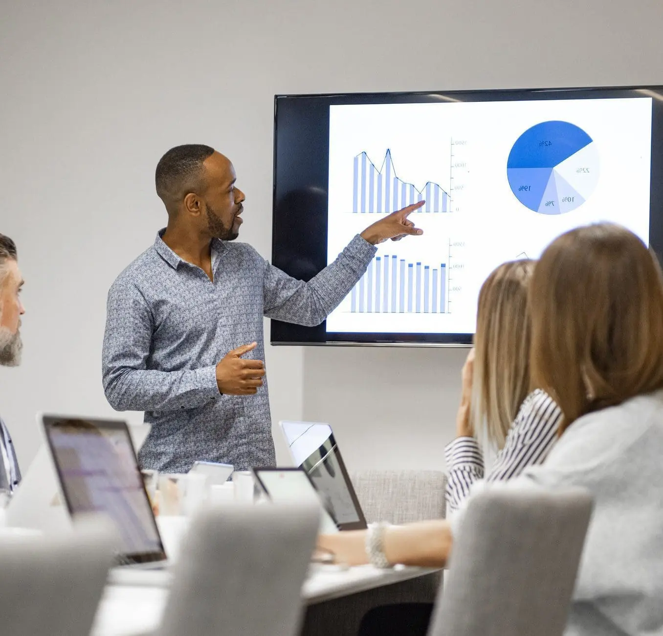 A Black man presents data visualizations on a large screen to a group of colleagues seated at a conference table.