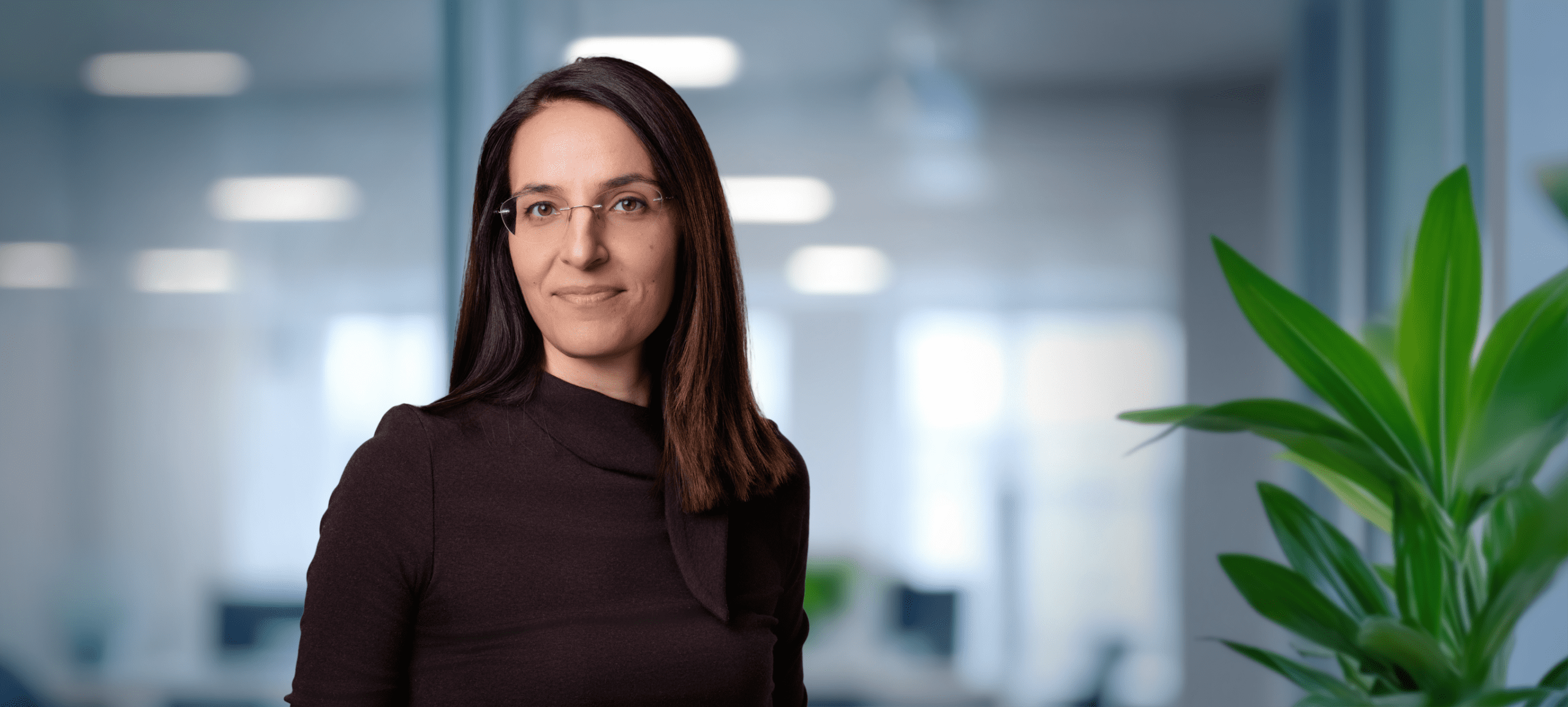 Adherent (formerly Compliance & Risks). Headshot of a smiling woman with long dark hair and rimless glasses, wearing a dark brown top, against a blurred office background with a green plant.