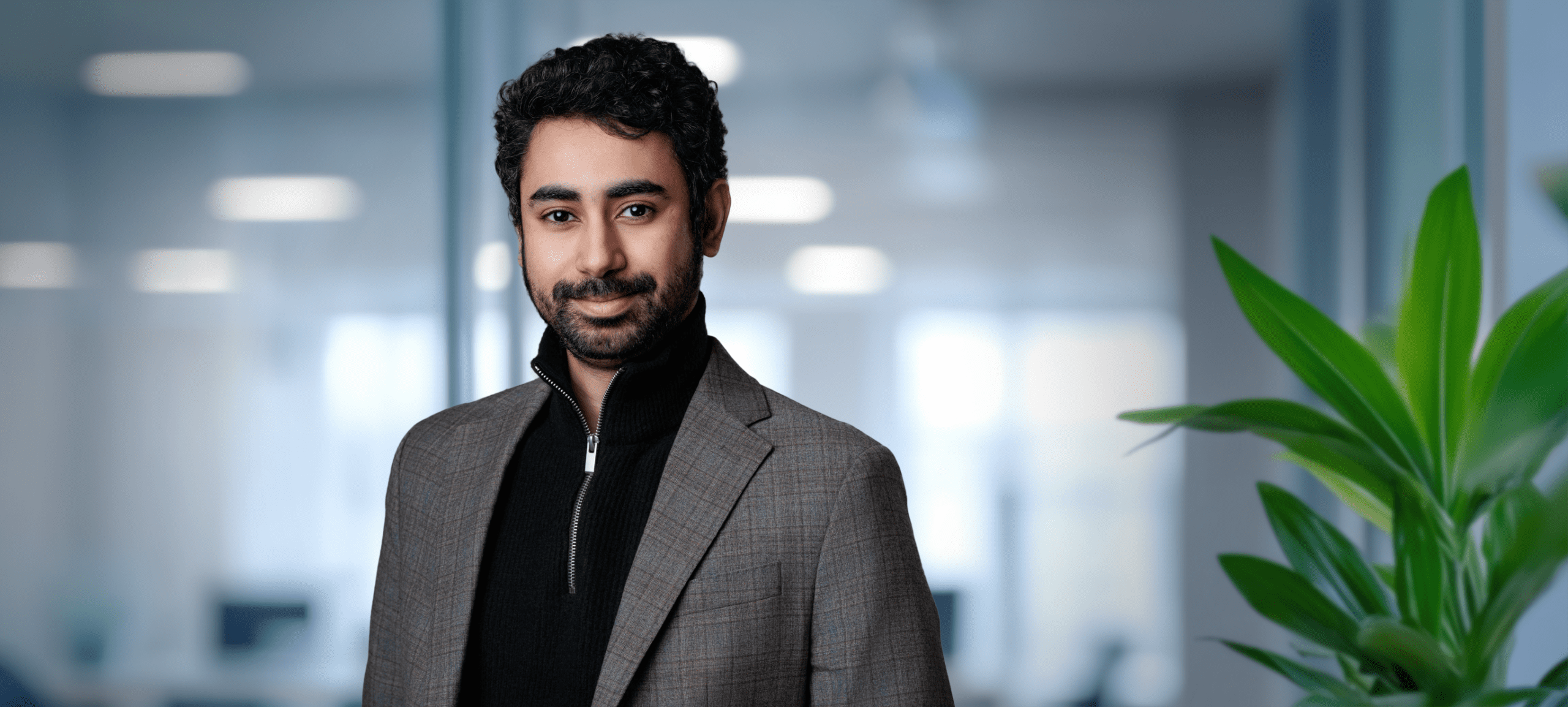 Adherent (formerly Compliance & Risks). A smiling man with dark curly hair and a beard, wearing a plaid blazer over a black quarter-zip sweater, in a modern office setting.