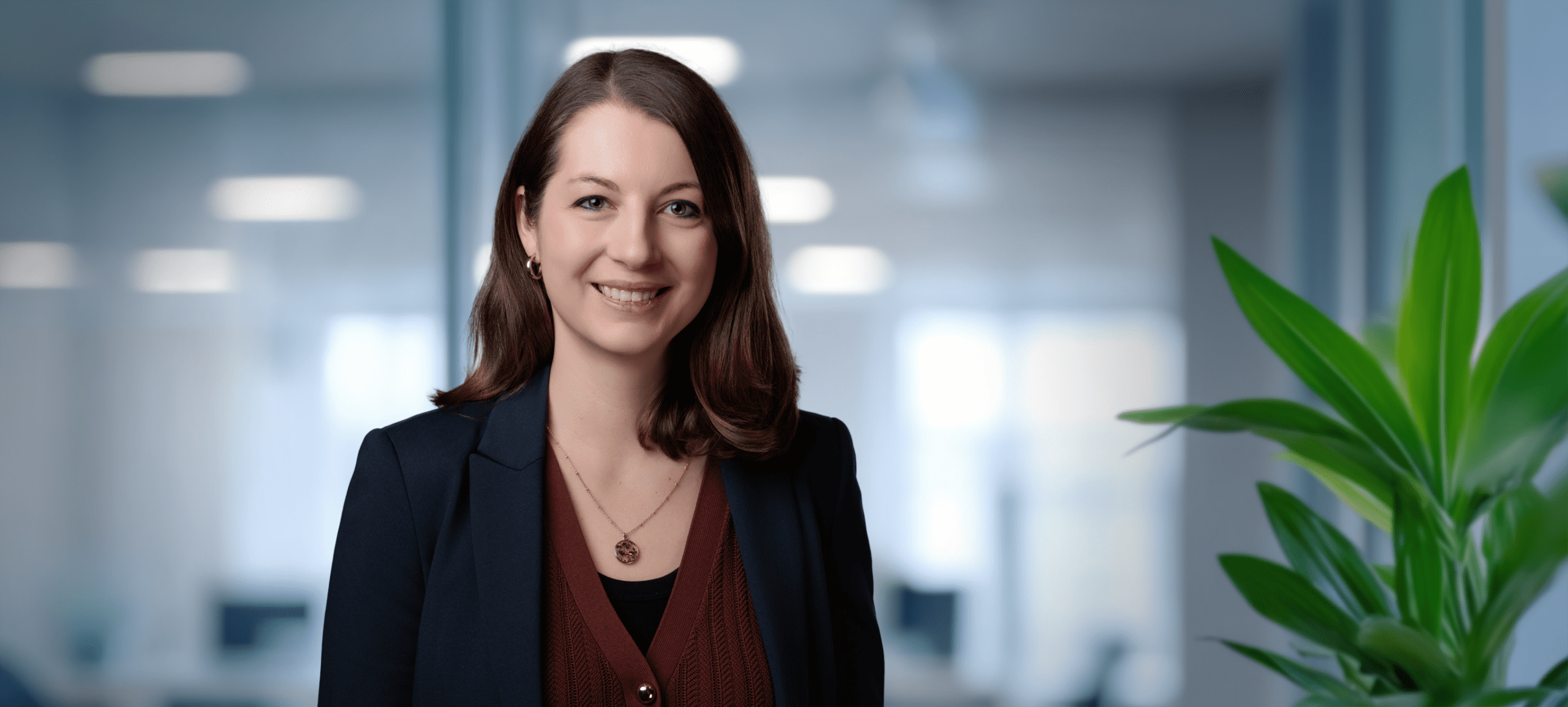 Headshot of a smiling woman with dark hair in a navy blazer, against a blurred office background with a green plant.