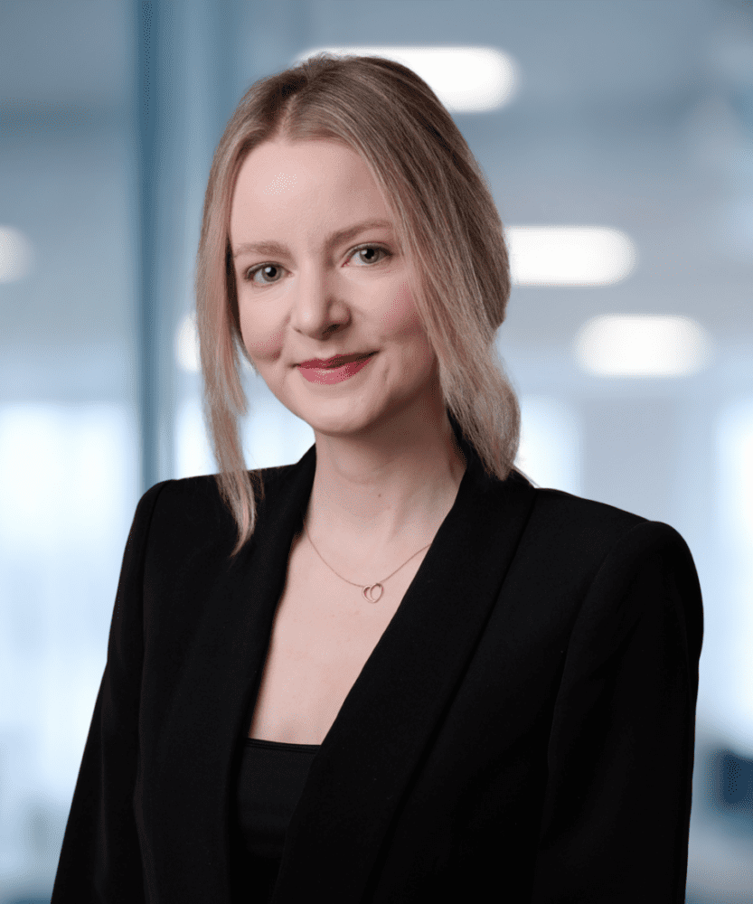 Headshot of a smiling woman with blonde hair wearing a black blazer and a gold necklace, in a blurred office background.