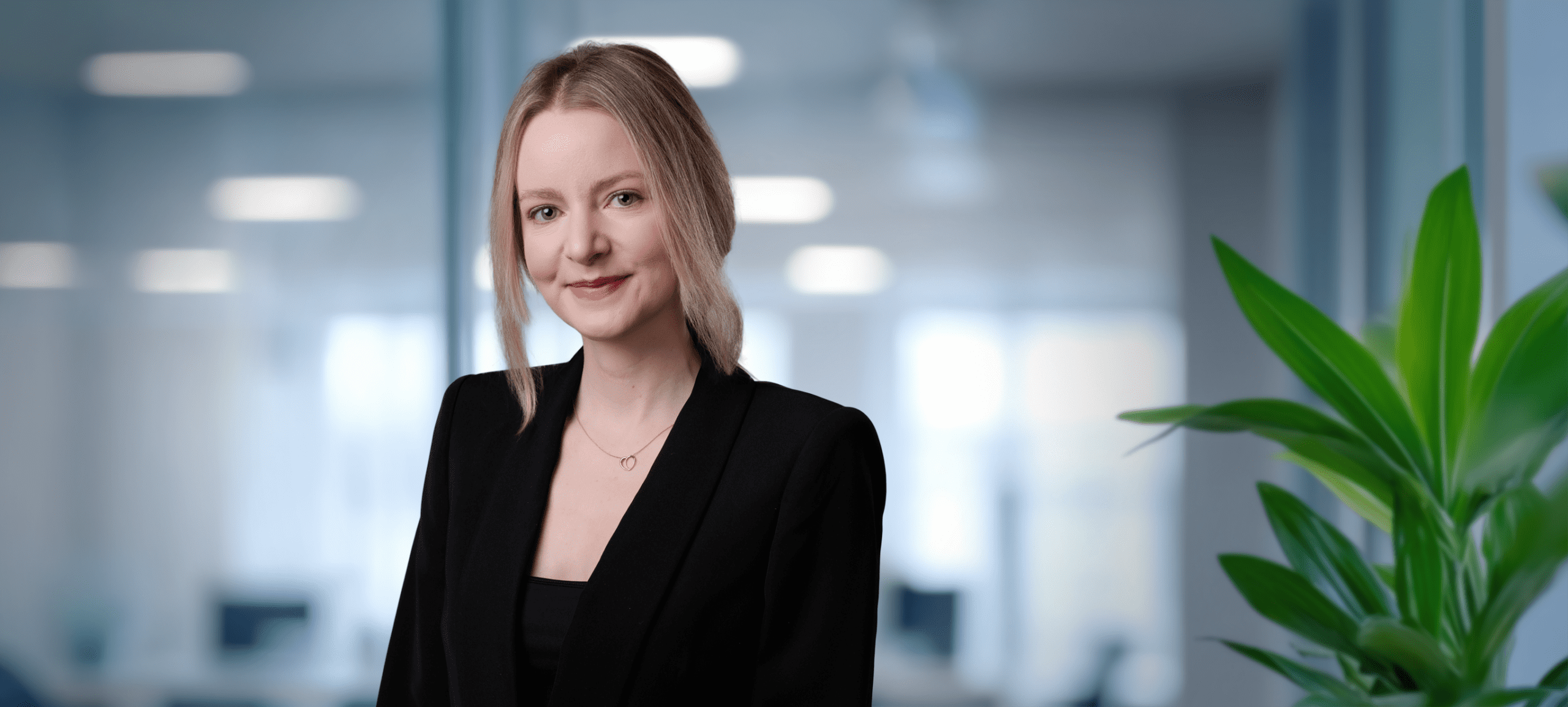 Adherent (formerly Compliance & Risks). Headshot of a smiling woman with blonde hair in a black blazer, wearing a gold necklace, against a blurred office background with a green plant.