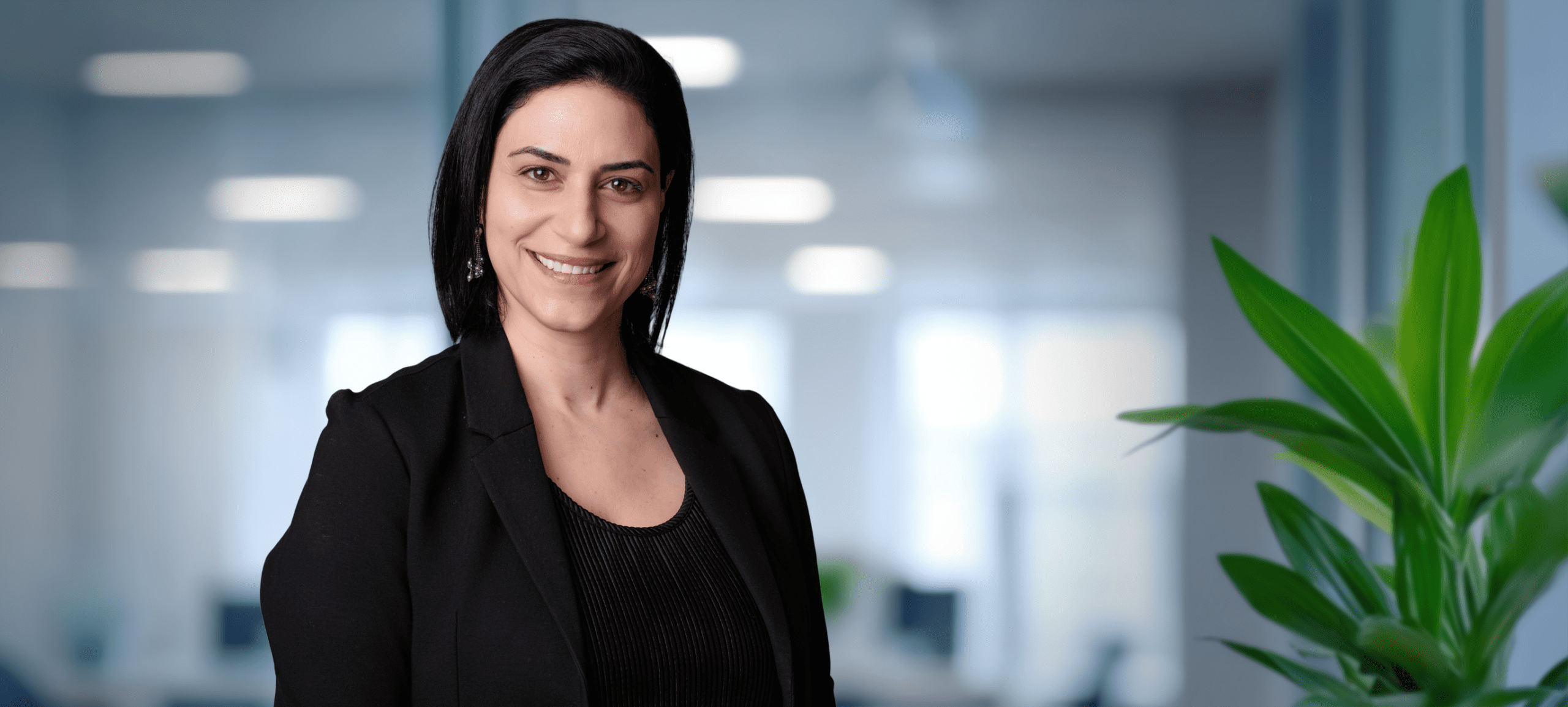 Adherent (formerly Compliance & Risks). Headshot of a smiling woman with dark hair in a black blazer, set against a blurred office background with a green plant.