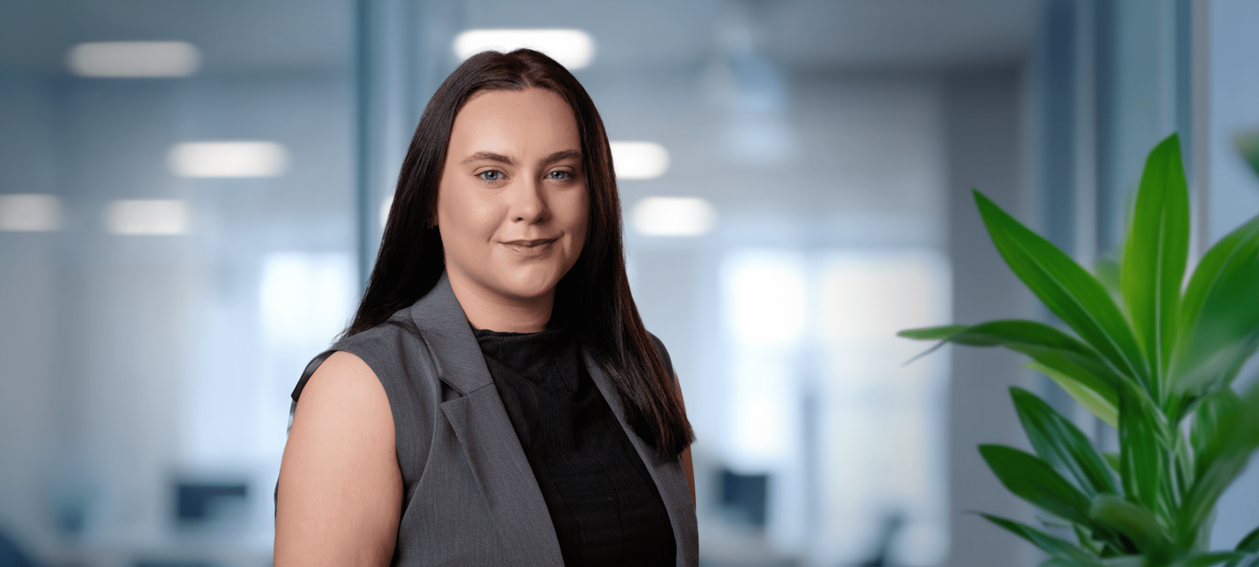 Adherent (formerly Compliance & Risks). A young woman with long dark hair and a grey vest smiles directly at the camera in a blurred office background with a green plant.