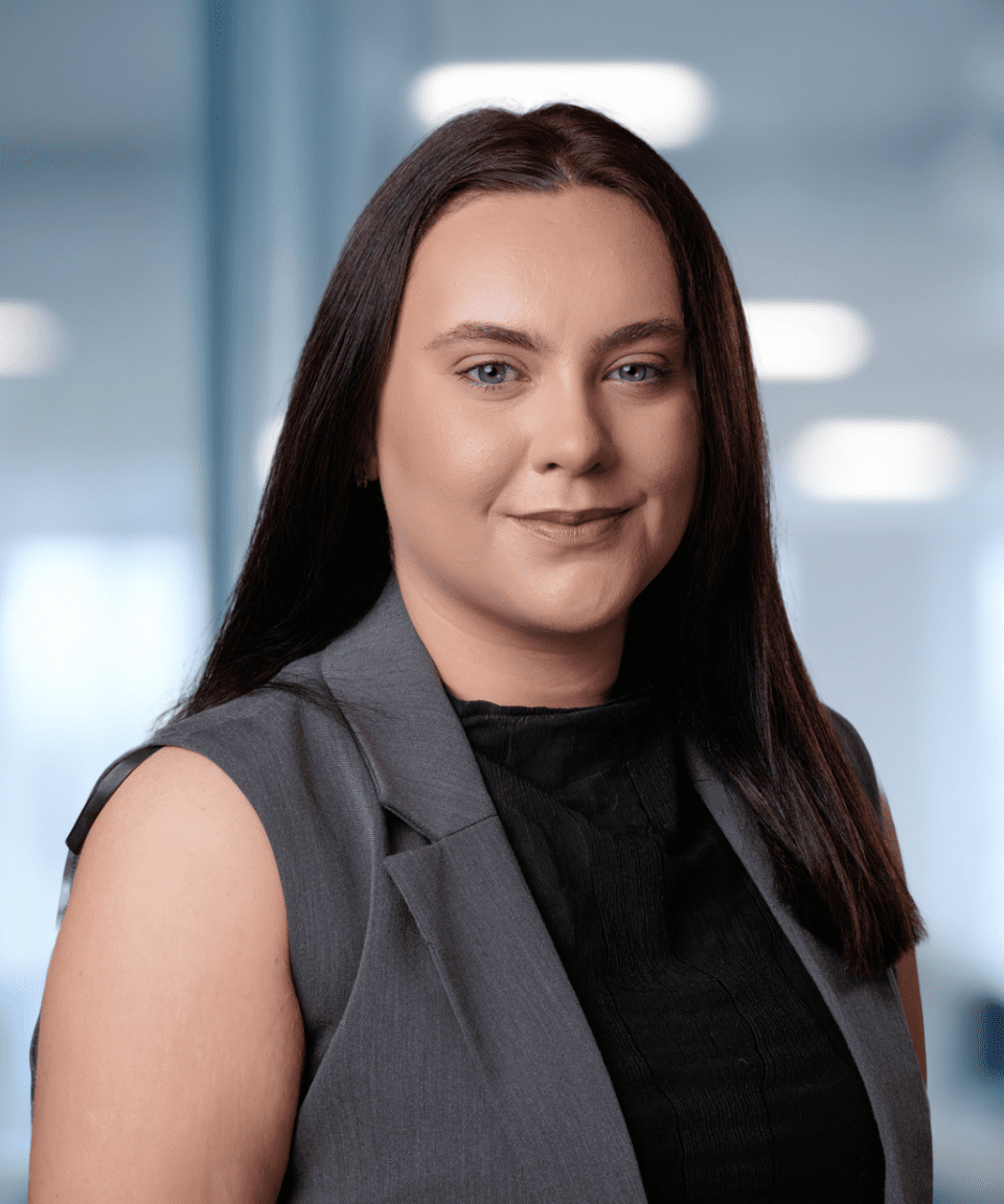 Adherent (formerly Compliance & Risks). A young woman with long dark hair and a grey vest smiles directly at the camera in a blurred office background with a green plant.