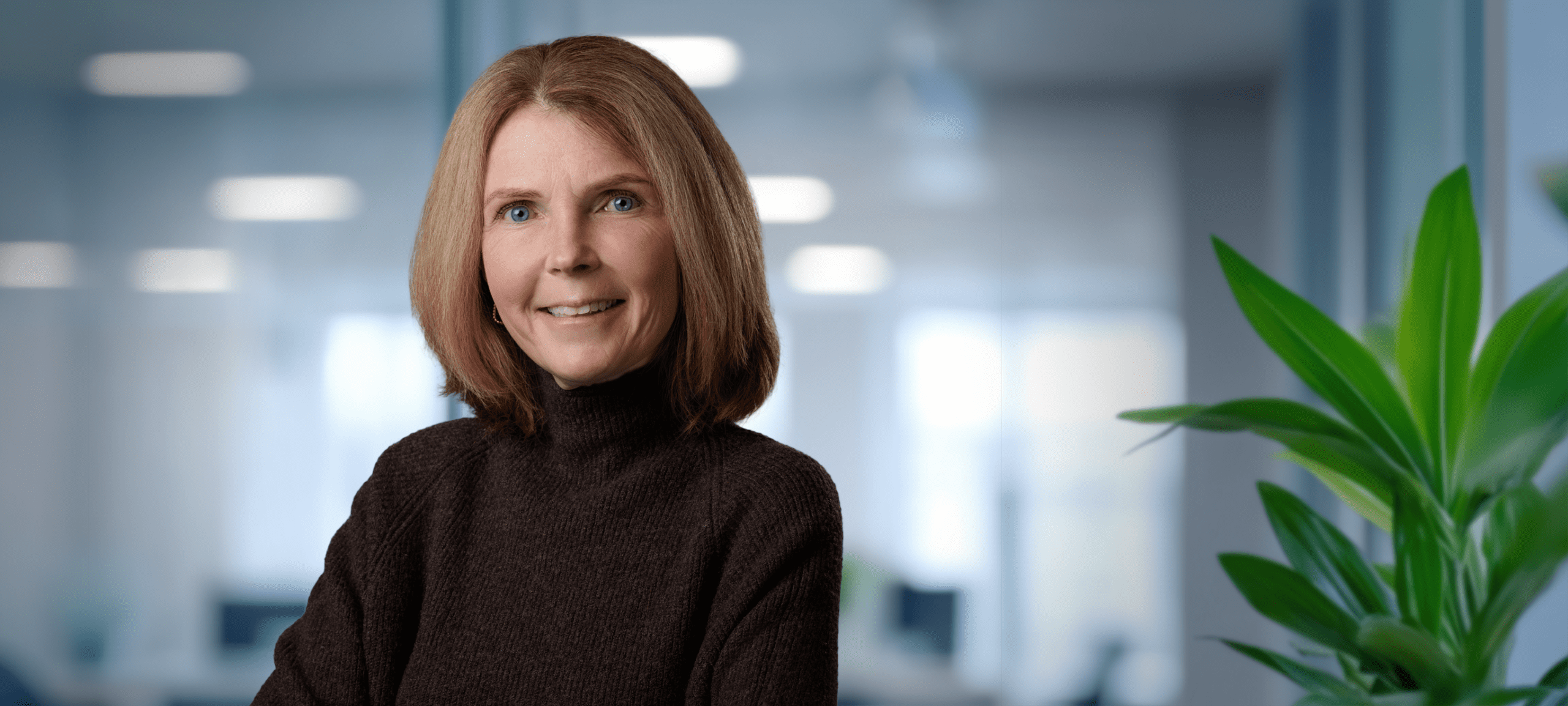 Adherent (formerly Compliance & Risks). Headshot of a smiling woman with light brown hair and blue eyes, wearing a dark brown turtleneck, against a blurred office background.