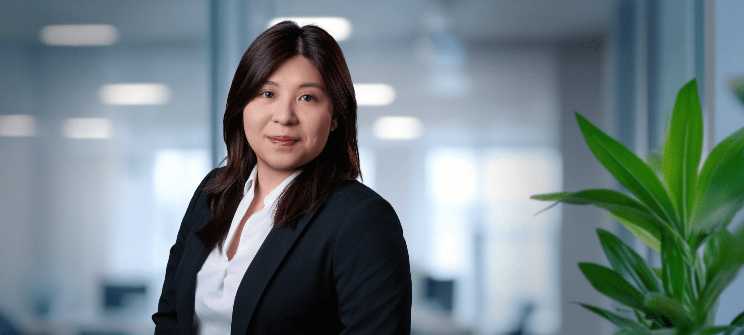 Adherent (formerly Compliance & Risks). Headshot of a smiling East Asian woman in a dark blazer and white shirt, set against a blurred modern office background with a green plant.